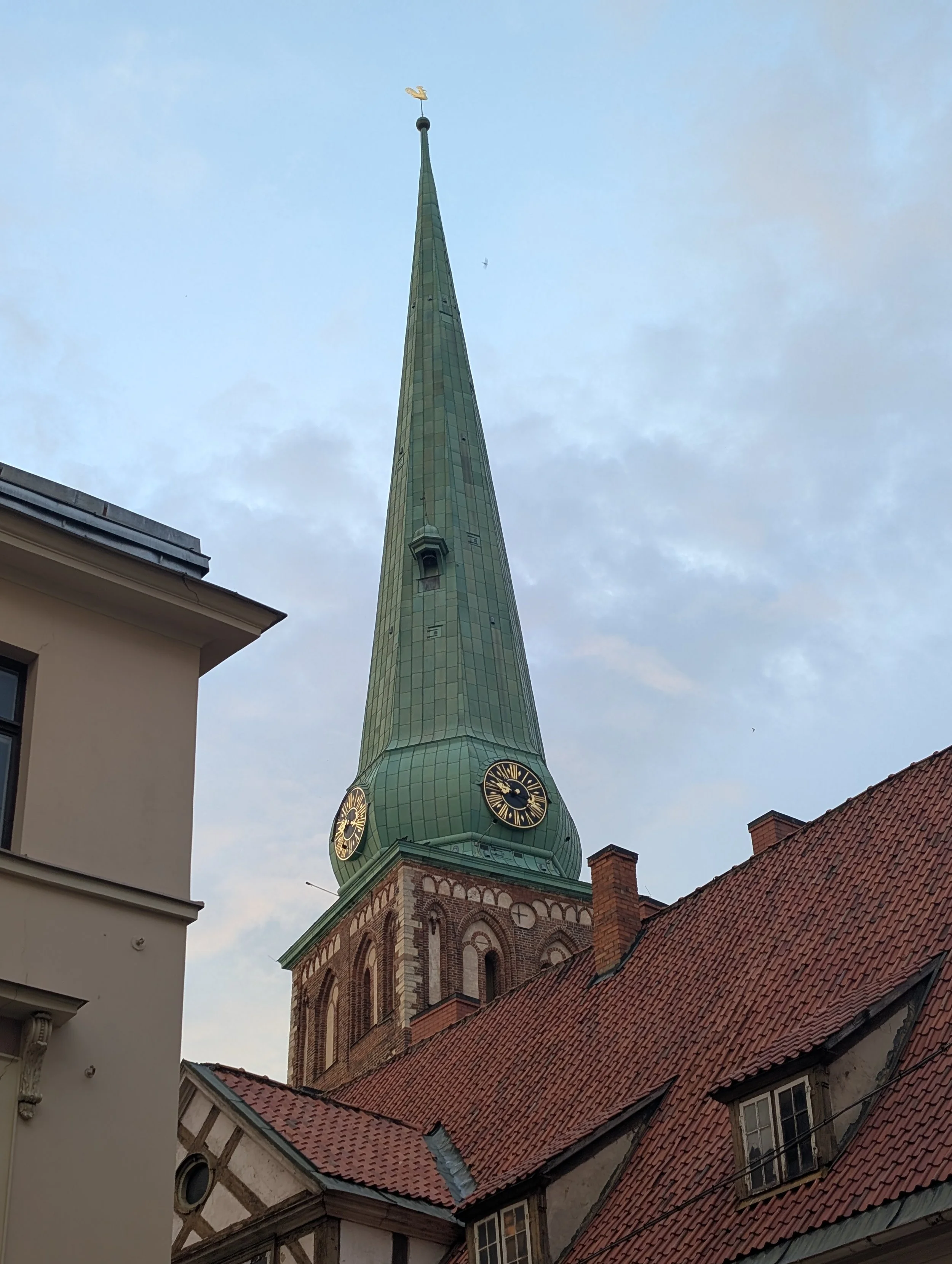 View of a church steeple with a clock, green copper roof, and a golden weather vane, surrounded by rooftops and part of a building with white walls and windows, under a cloudy sky.