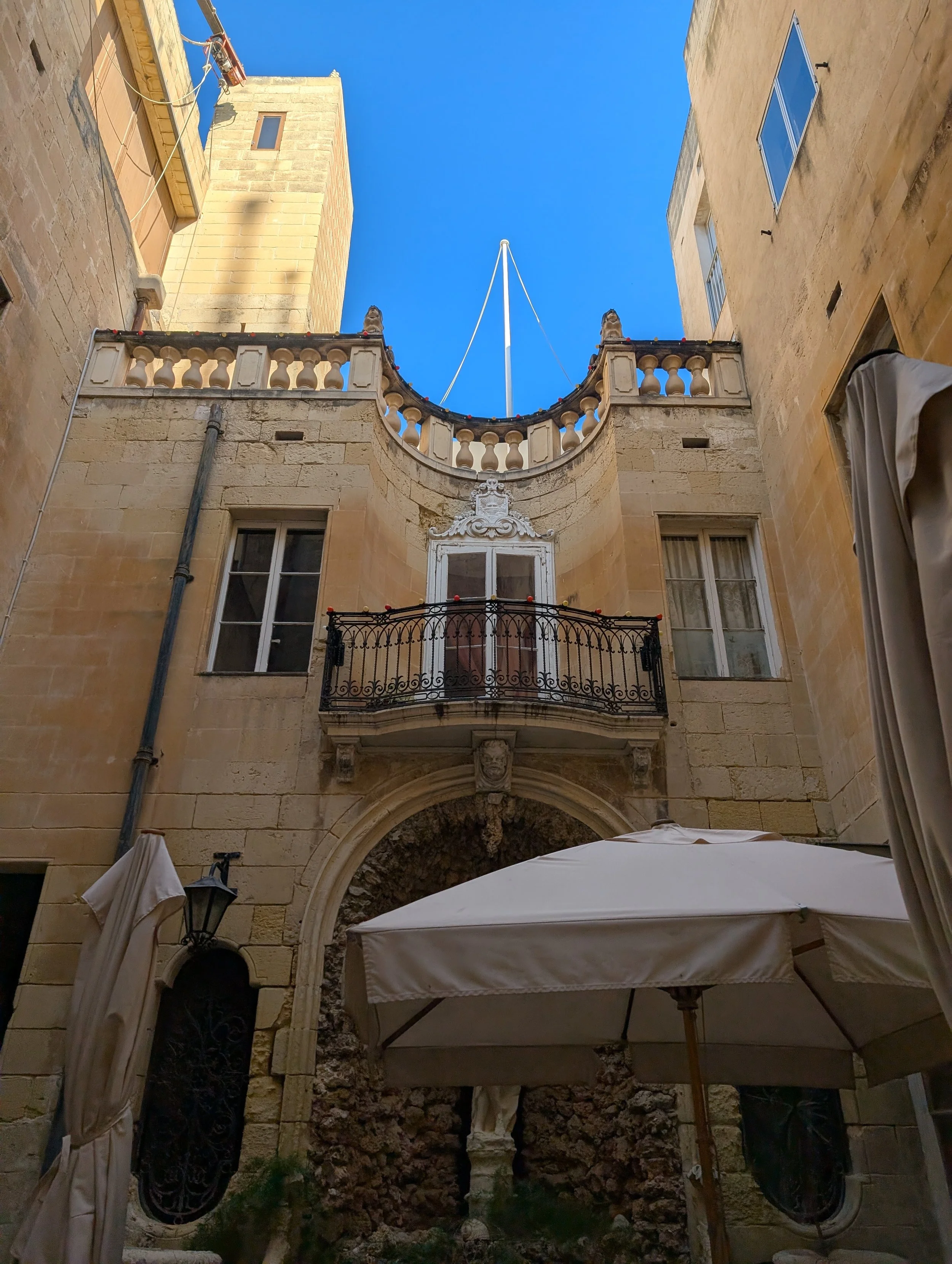 View of a narrow courtyard surrounded by historic stone buildings with arched doorways, a balcony with ornate railing, closed windows, and umbrellas shading an outdoor seating area under a blue sky.