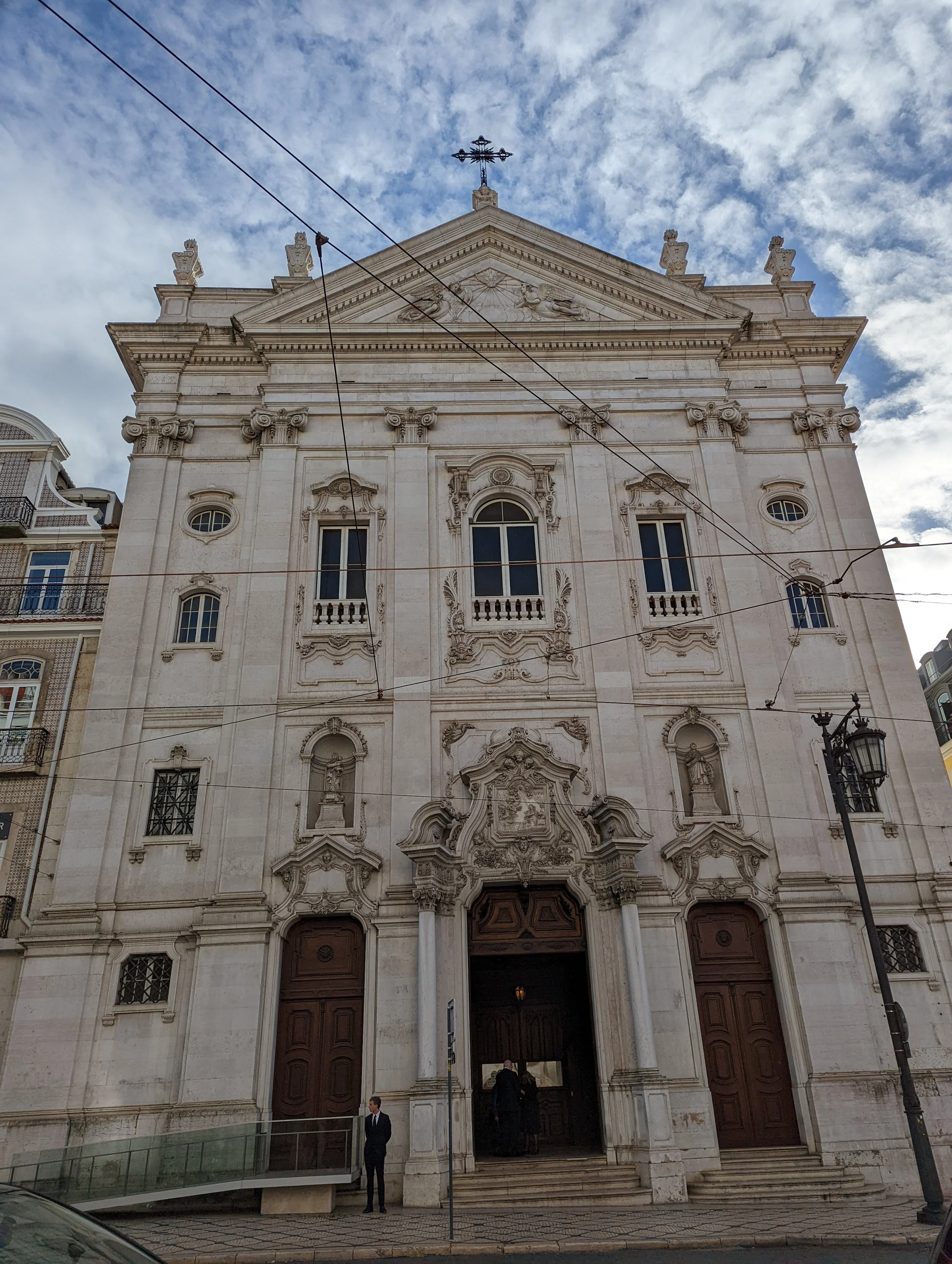 Facade of a historic stone church with ornate architectural details, large wooden doors, and a cross on the roof top, under a partly cloudy sky.