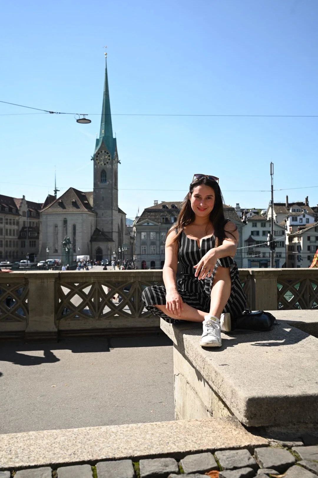 A woman sitting on a stone ledge in front of a historic city square with a church and clock tower in the background on a sunny day.
