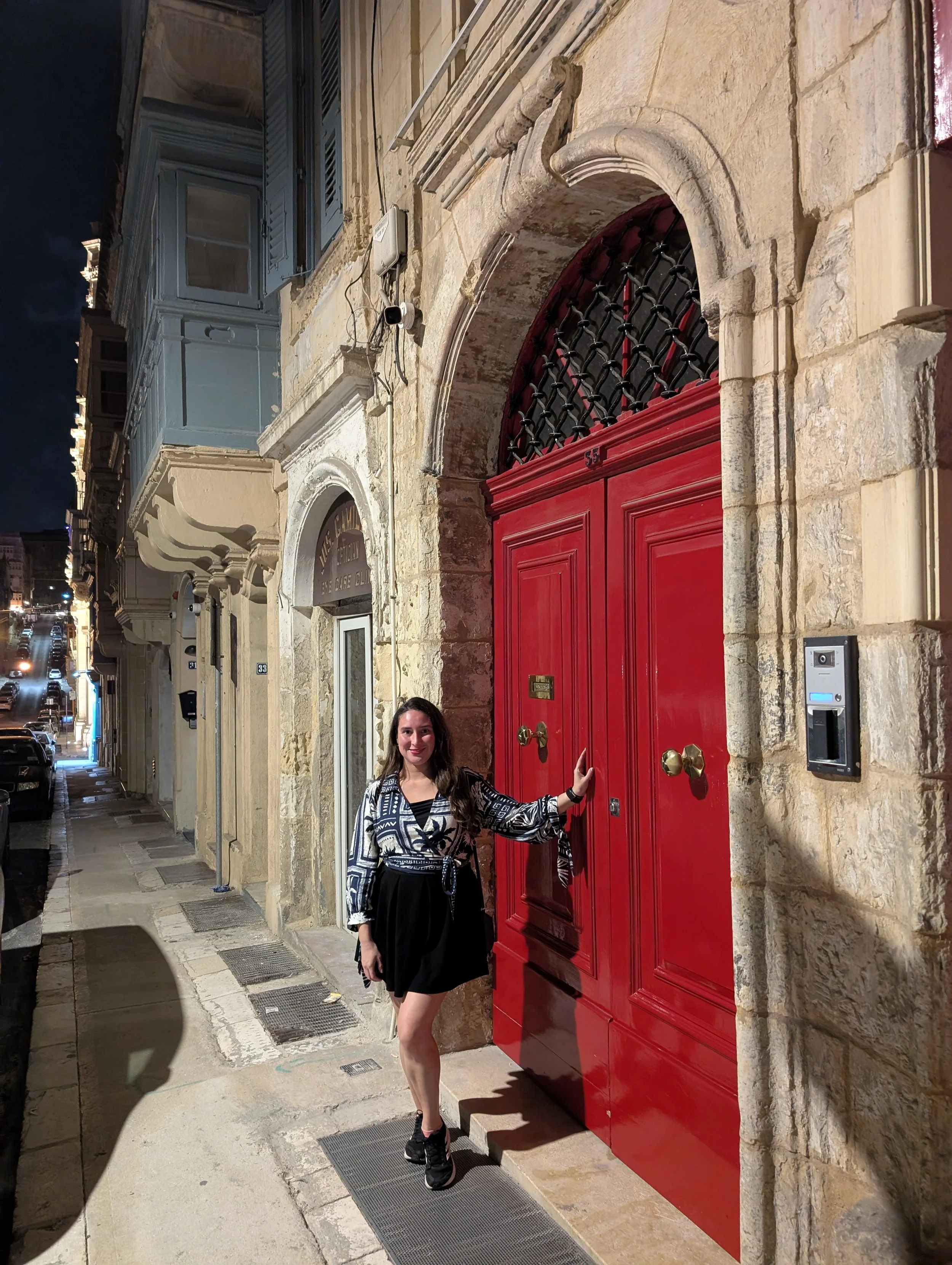 A woman in a black skirt and patterned blouse stands on a sidewalk in front of a red door with a decorative iron grate on top, holding the door handle. The building is made of stone with an arched entrance, and there are other buildings and parked ca