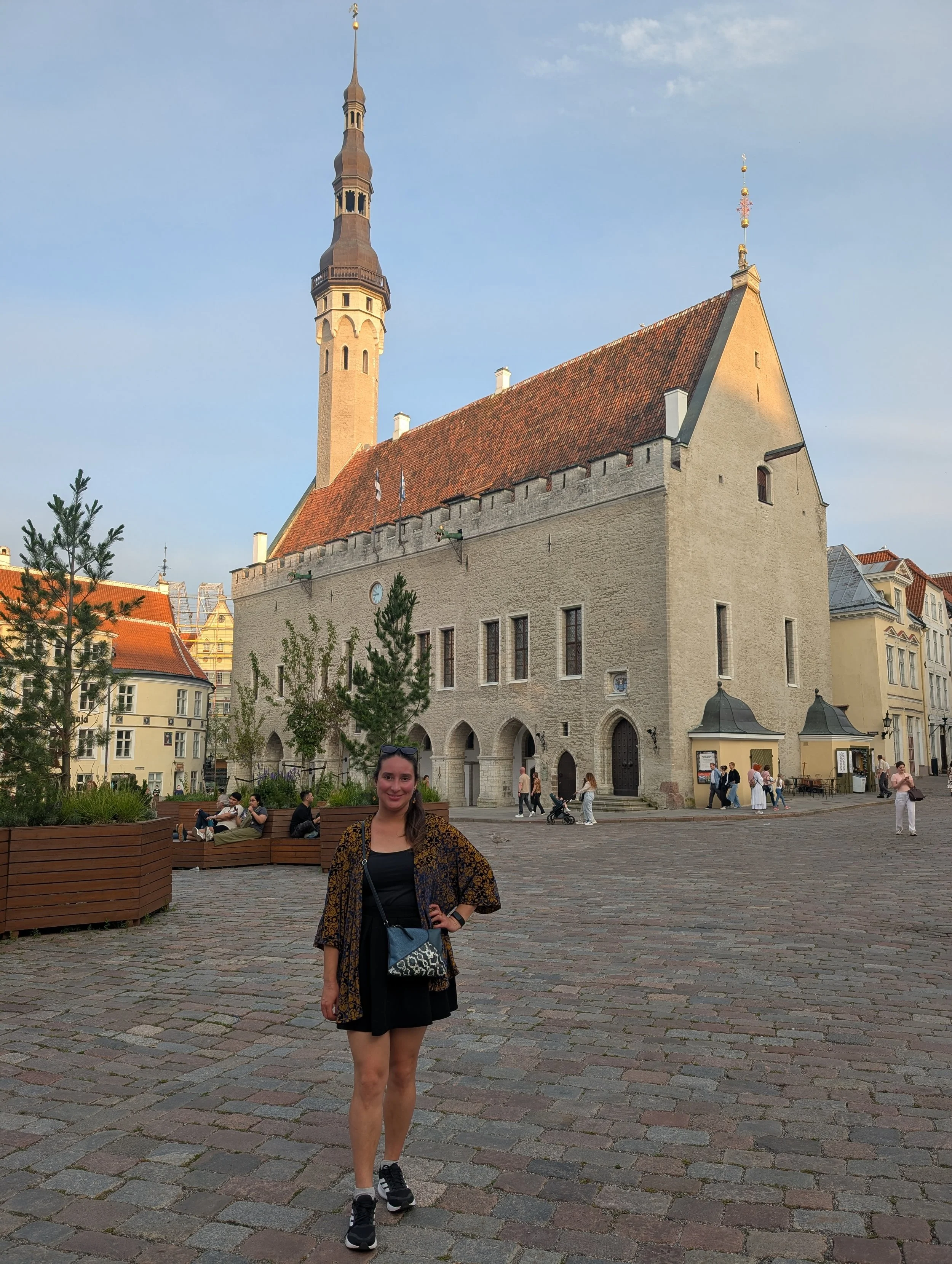 A young woman with long hair, wearing a black dress, patterned jacket, and sneakers, standing in a cobblestone square with historic buildings and a church with a tall spire in the background, during daytime.