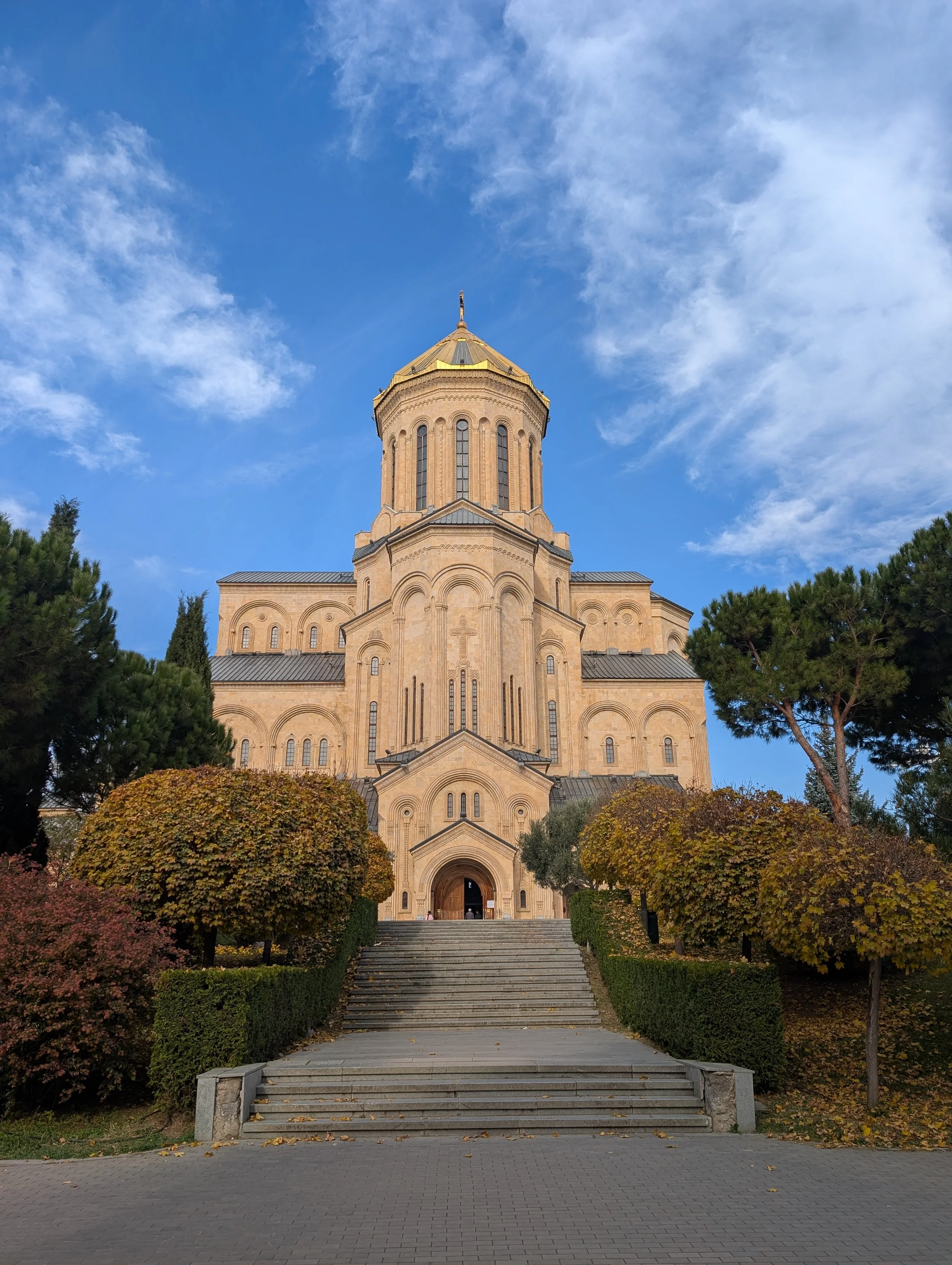 Tall church with a golden dome at the top, surrounded by trees with autumn leaves, blue sky with wispy clouds, stone stairs leading up to the entrance.