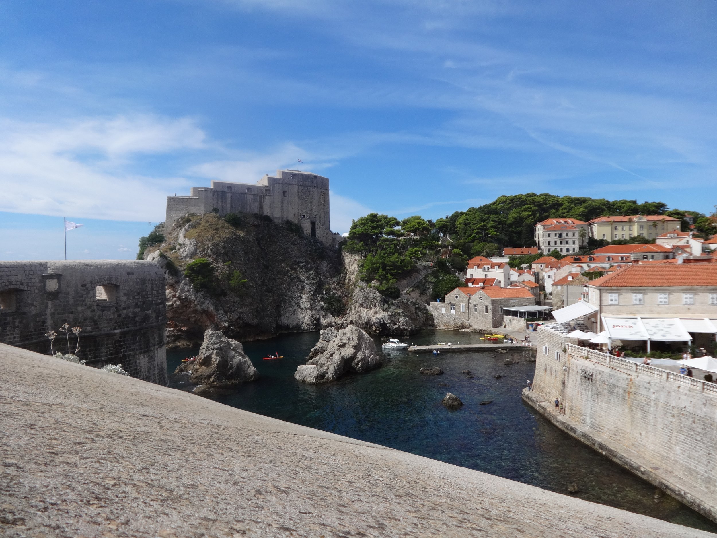 Historic stone fortress on a rocky hill overlooking a harbor with boats, surrounded by buildings with red-tiled roofs and lush green trees under a partly cloudy sky.