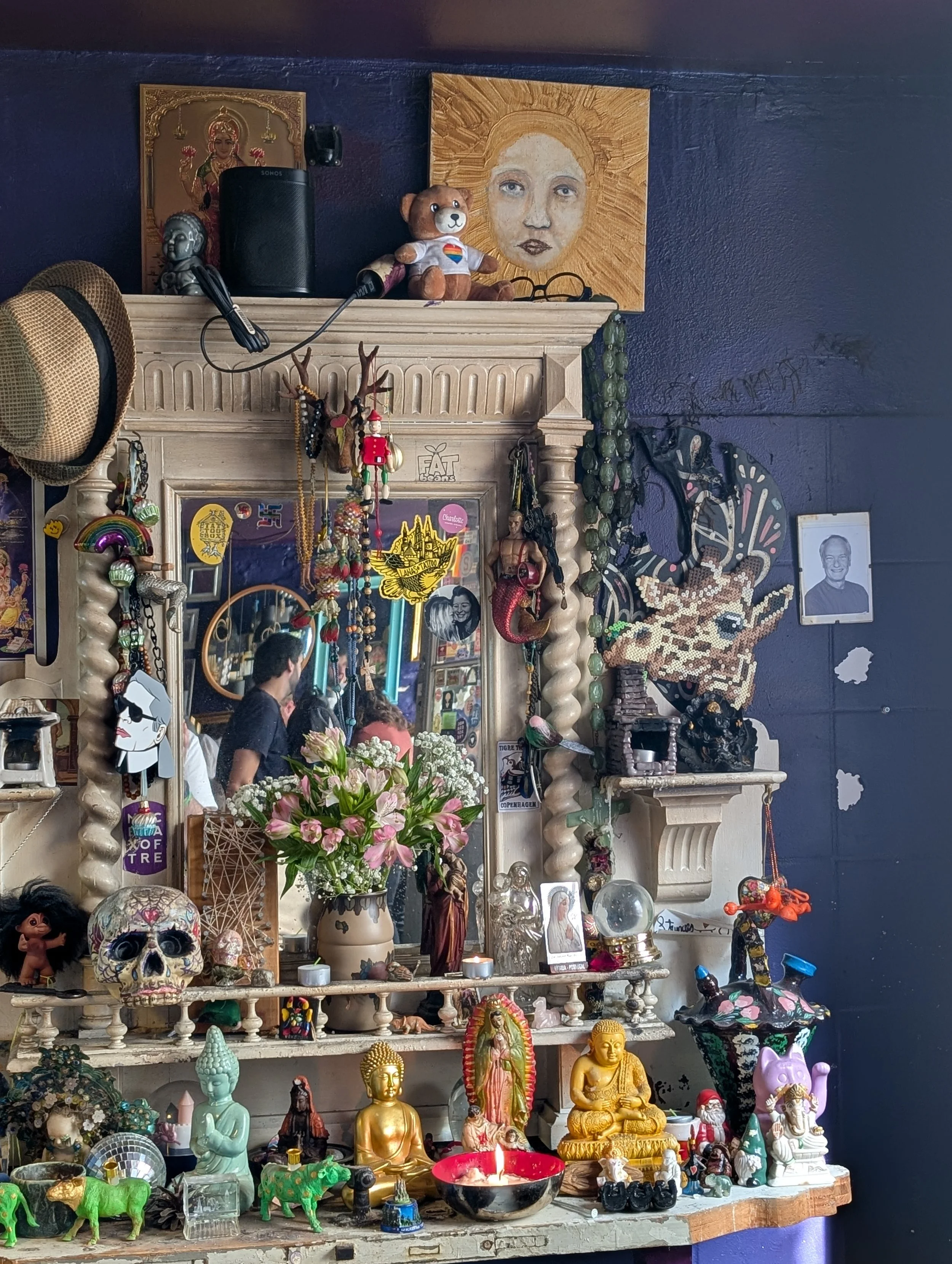 Eclectic altar with religious statues, skull figurines, candles, and various decorative objects against a dark wall.