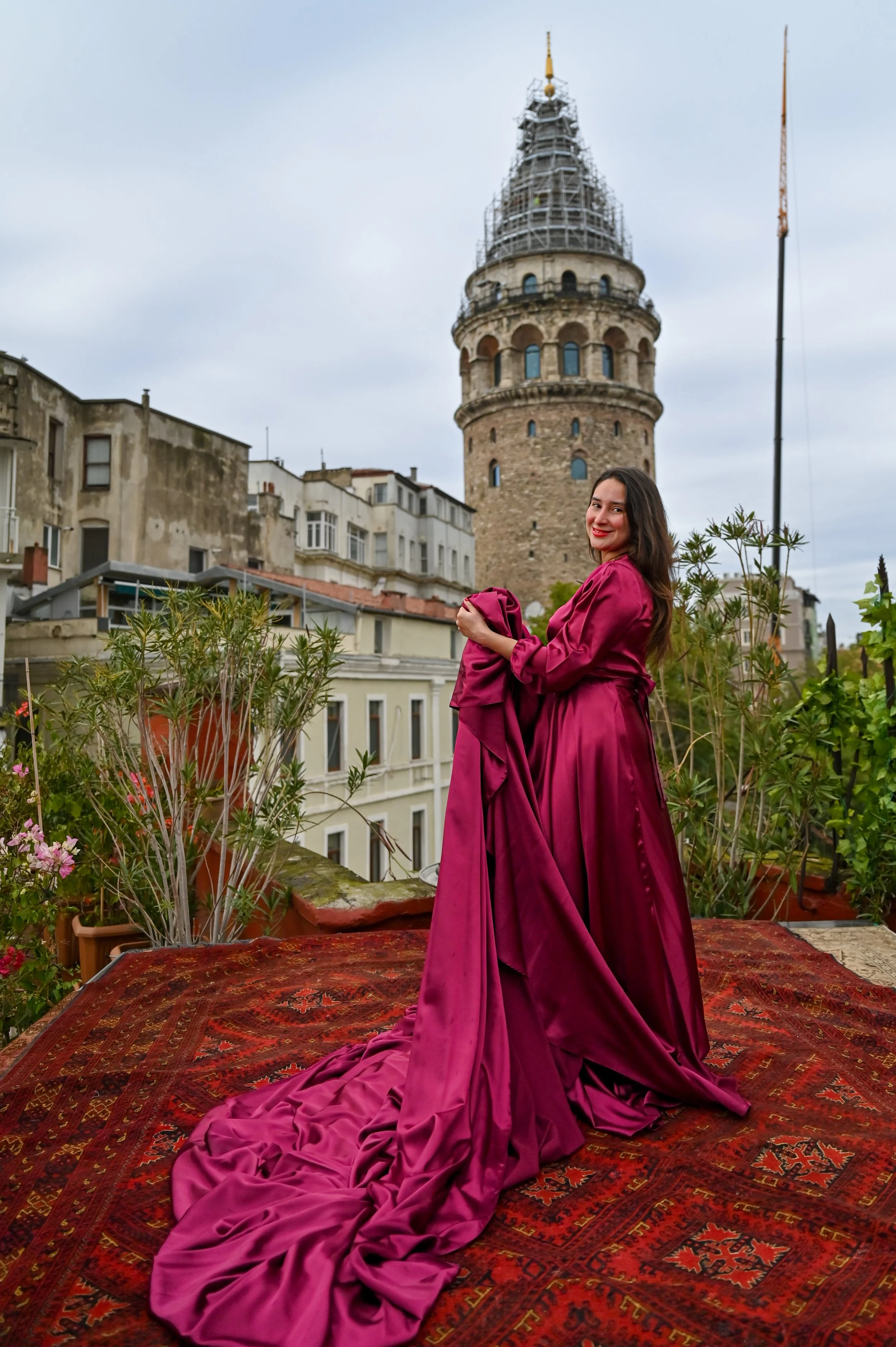 A woman in a long pink gown on a rooftop garden with plants and flowers, with historic buildings and the Galata Tower in Istanbul in the background.