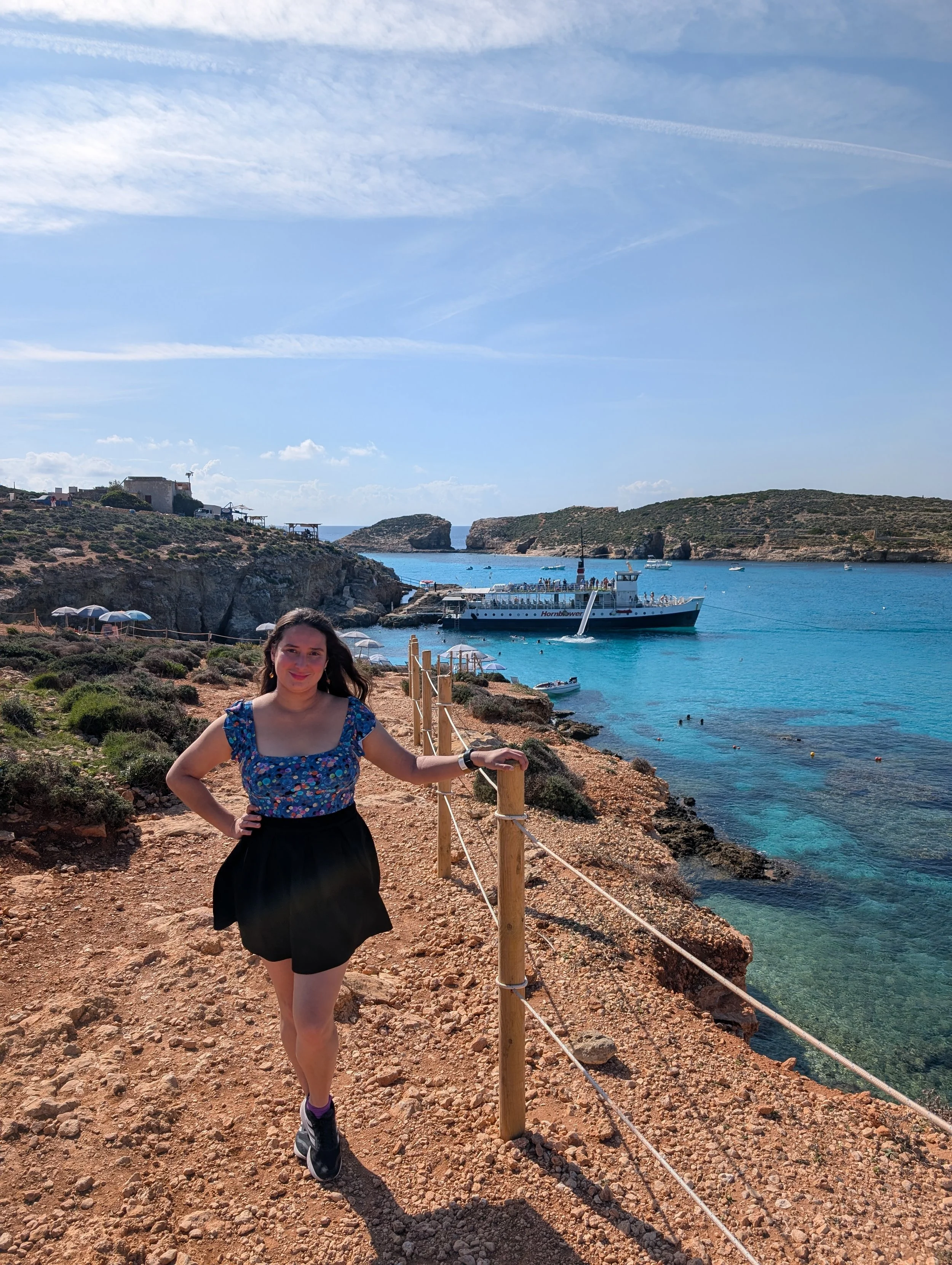 A woman with dark hair, wearing a blue floral top, black skirt, and sneakers, standing on a rocky pathway by the sea, with a fence and a boat in the background, under a partly cloudy sky.