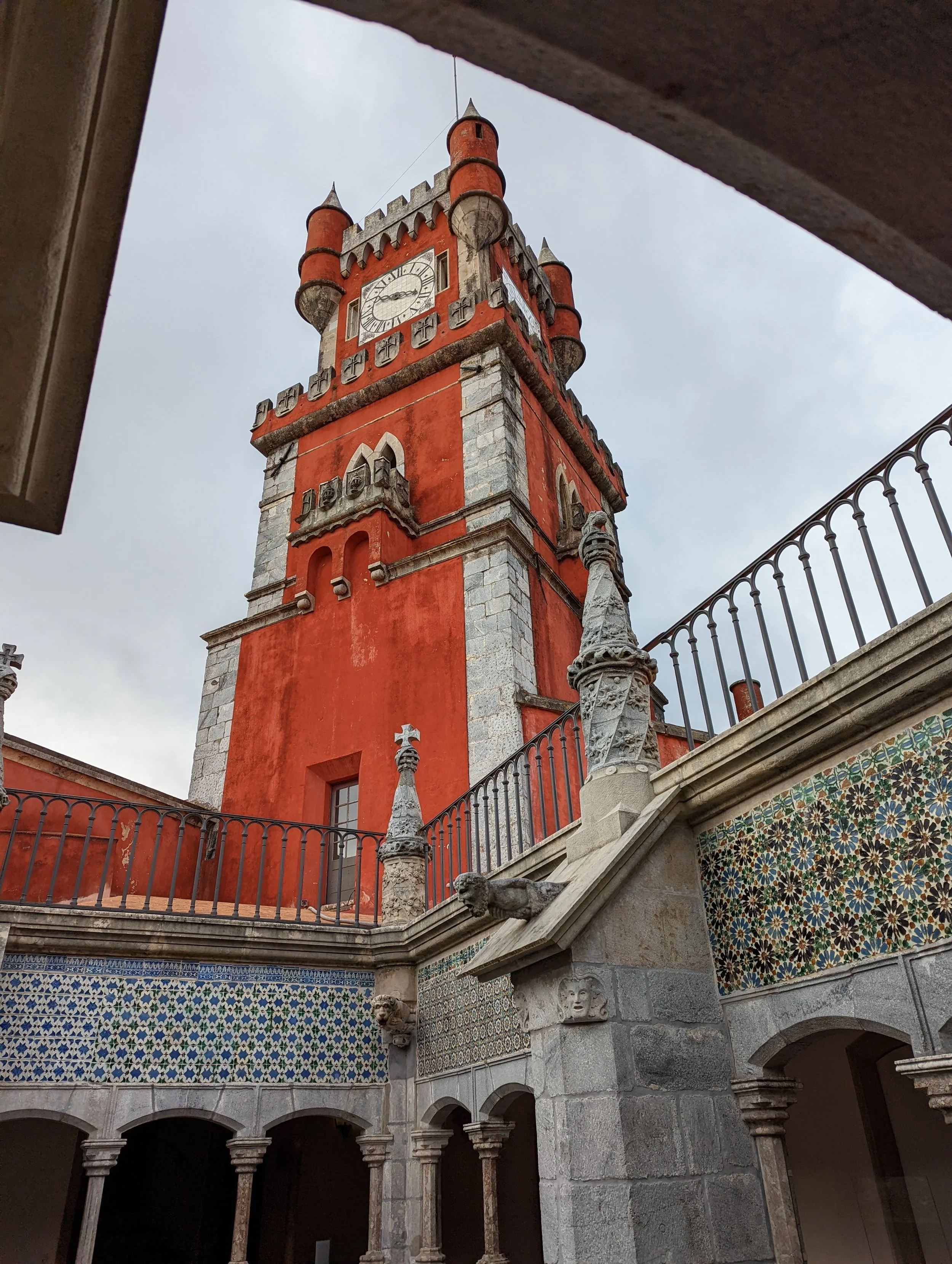 A historic clock tower with a red facade and decorative turrets, viewed from below, surrounded by an ornate stone balcony with colorful geometric tile work and a railing. The sky in the background is cloudy.