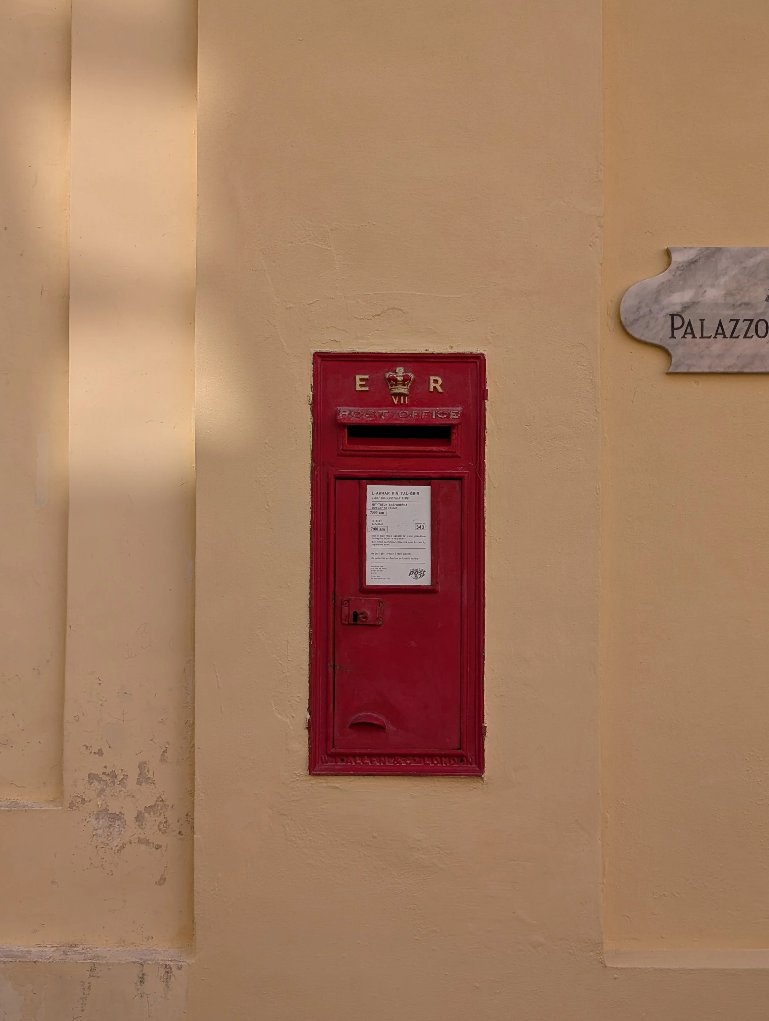 A red postal mailbox labeled 'E R' with a crown symbol above the letters, mounted on a beige wall next to a sign that reads 'PALAZZO' on a marble plaque.