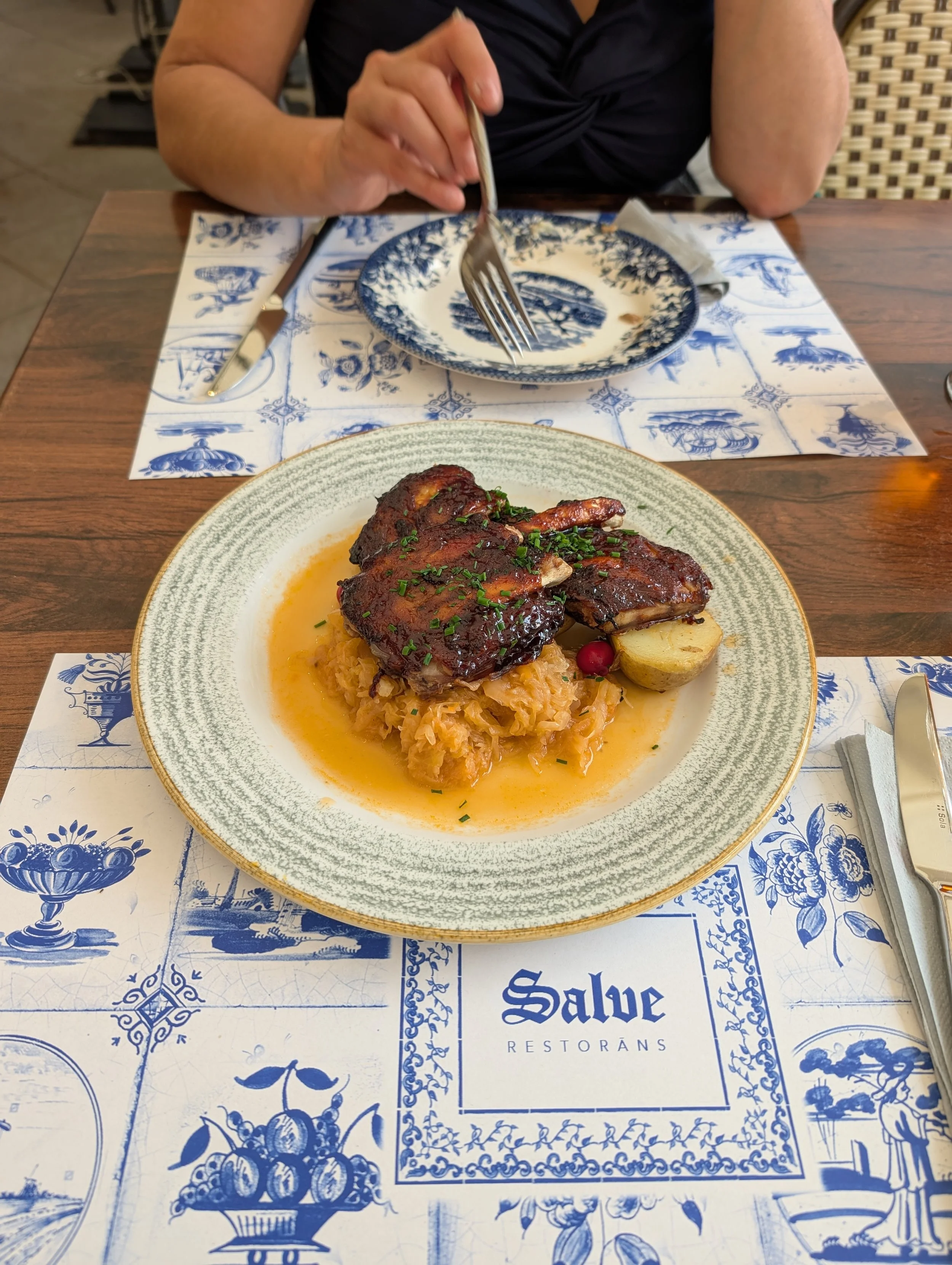A plate of cooked steak with gravy, mashed potatoes, and garnish, on a decorative plate on a table with blue and white patterned paper and utensils.