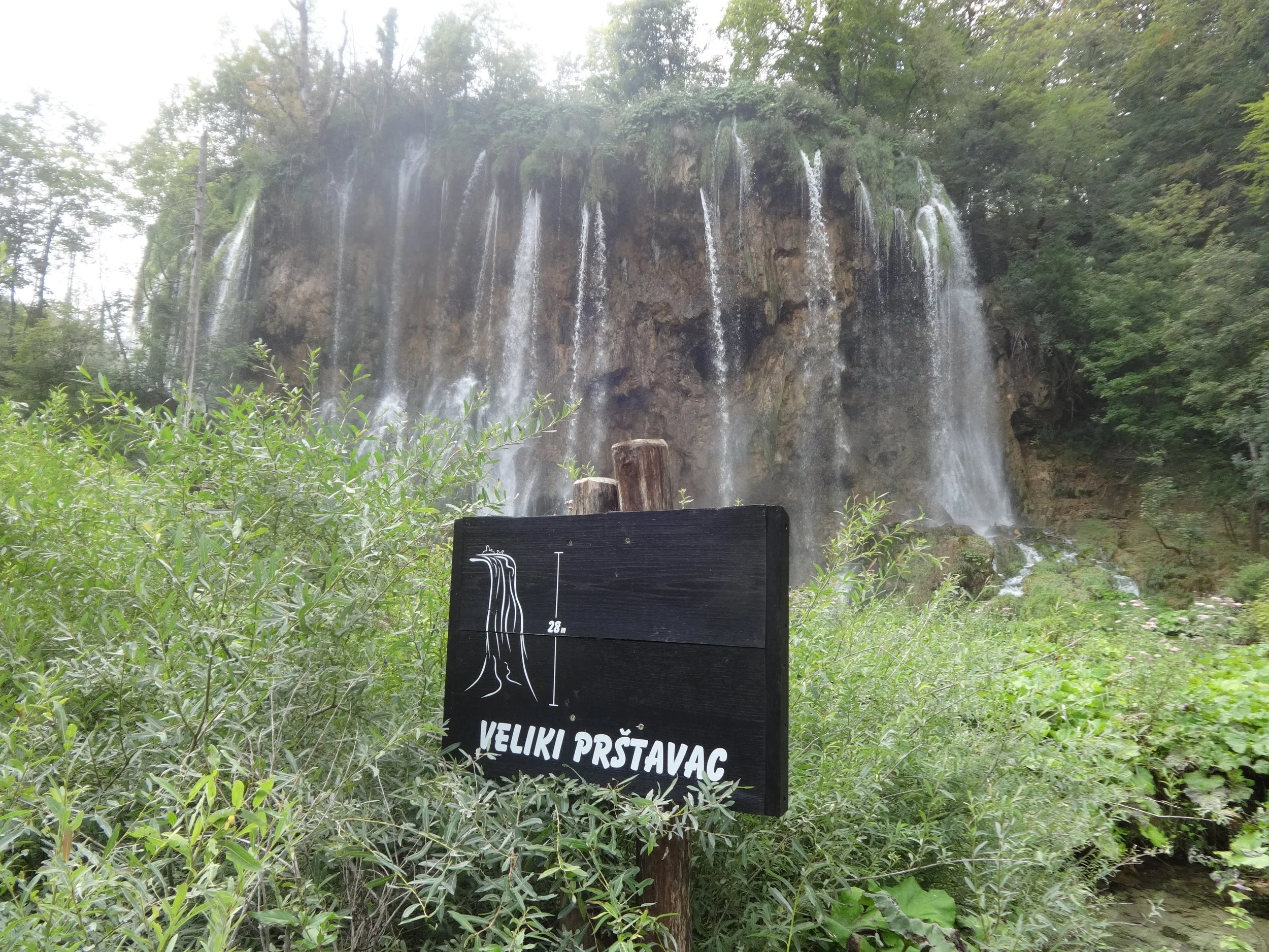 A waterfall cascading down a rocky cliff surrounded by green trees and bushes, with a sign in front indicating the height of the waterfall.