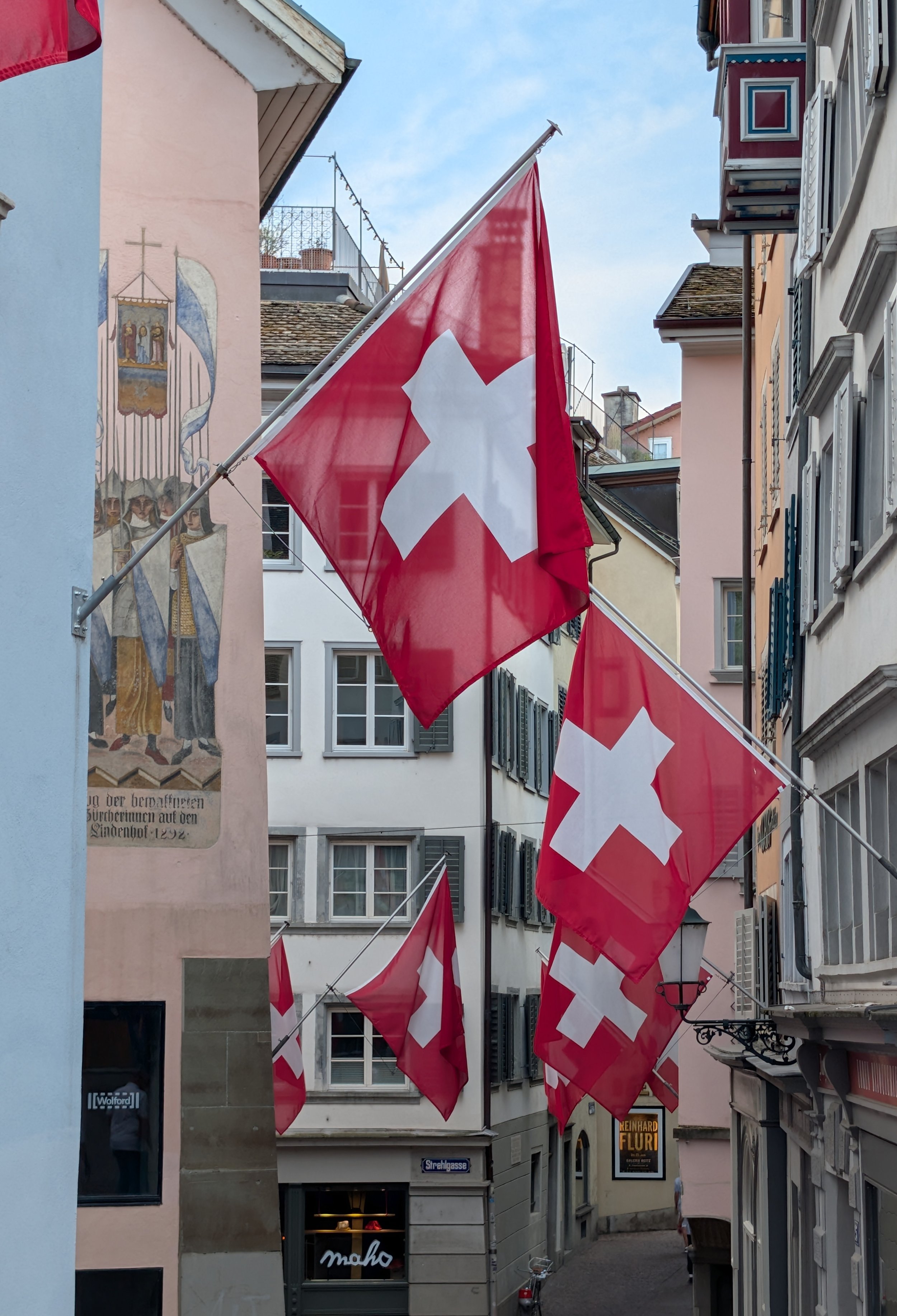 Swiss flags hanging along a narrow street in a European city, with pastel-colored buildings and a mural on the wall to the left.