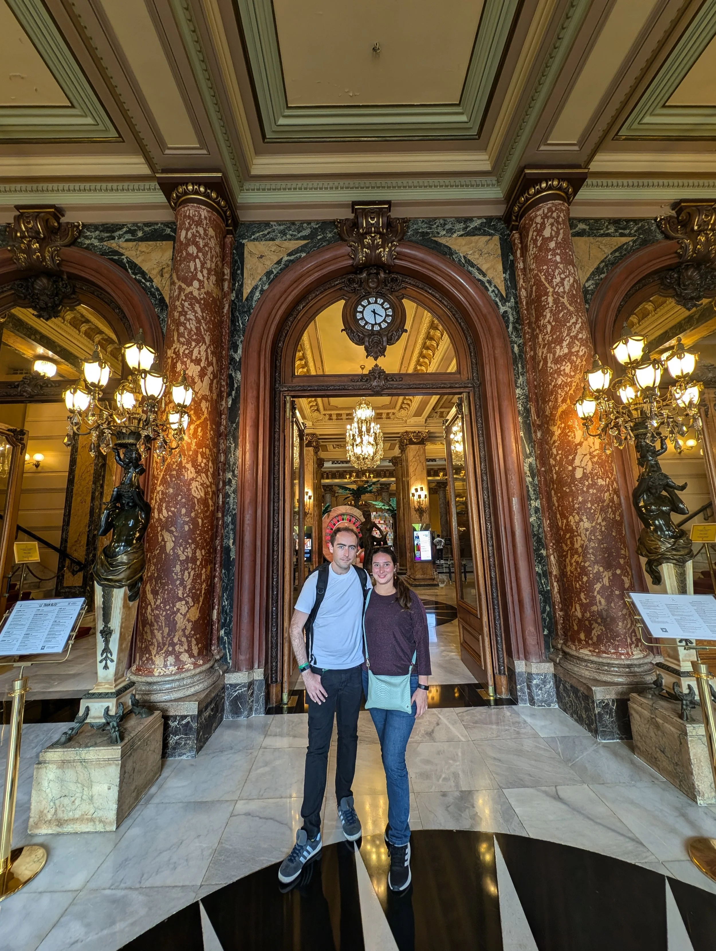 Two people standing inside a luxurious building with ornate columns, chandeliers, and classical architectural details.