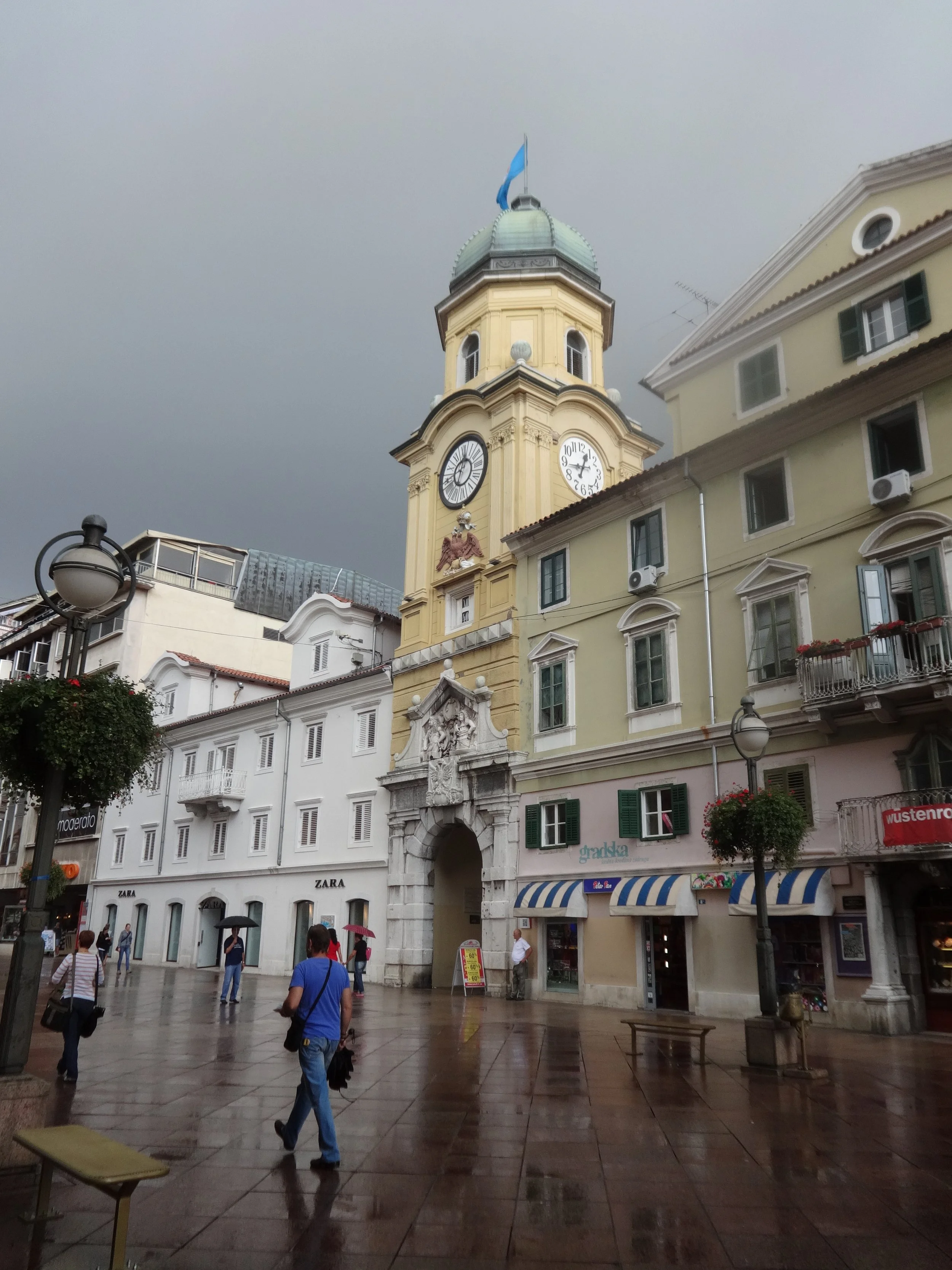 A city square with a yellow clock tower, a building with retail stores including Zara, and people walking on a wet pavement under cloudy skies.