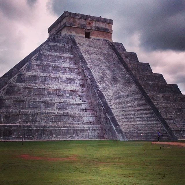 A large stepped pyramid made of stone, with a small structure on top, set against a cloudy sky with green grass in the foreground.
