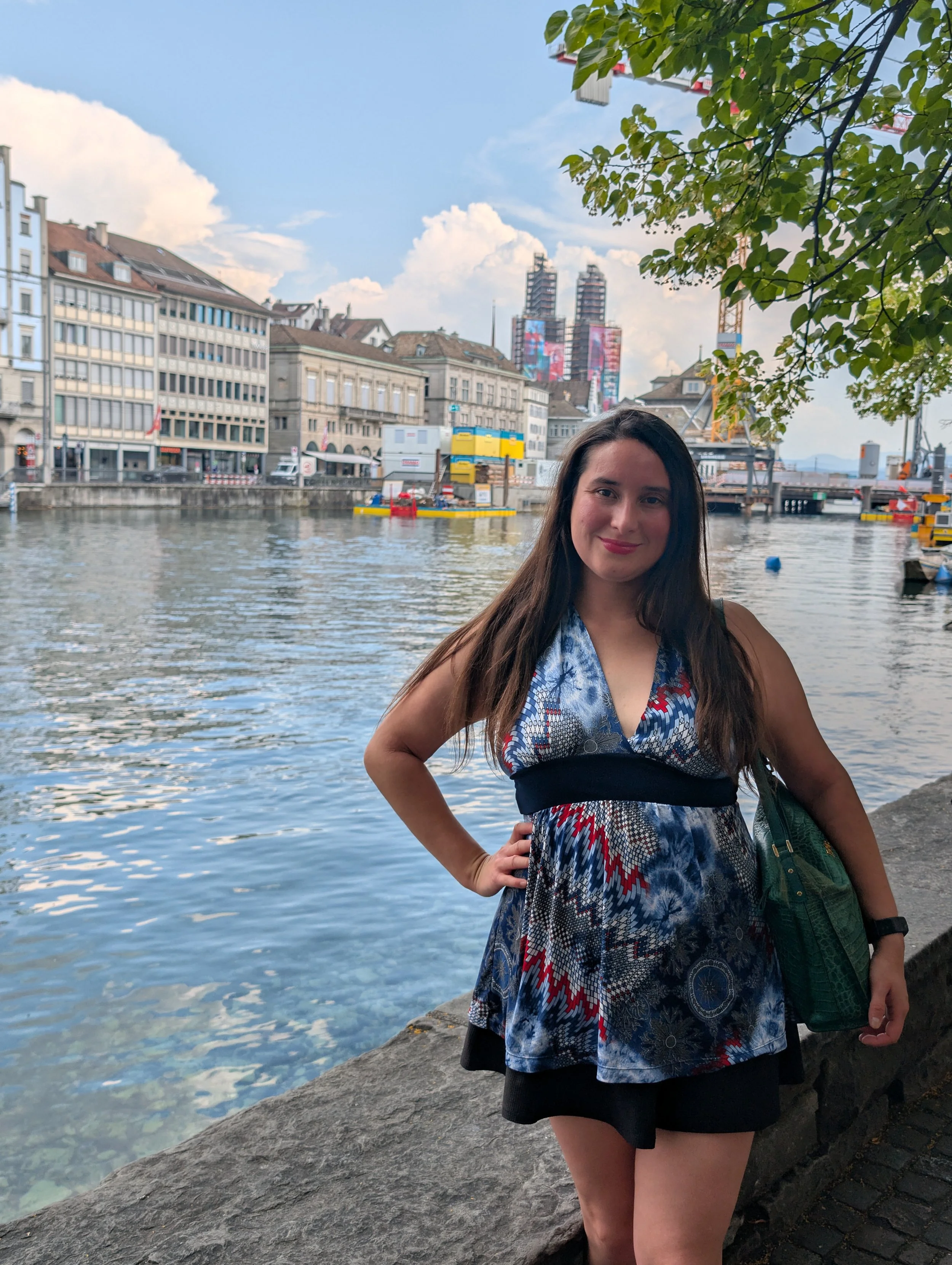 A woman with long dark hair standing on a riverbank with city buildings and a bridge in the background, wearing a colorful patterned dress and carrying a green bag.