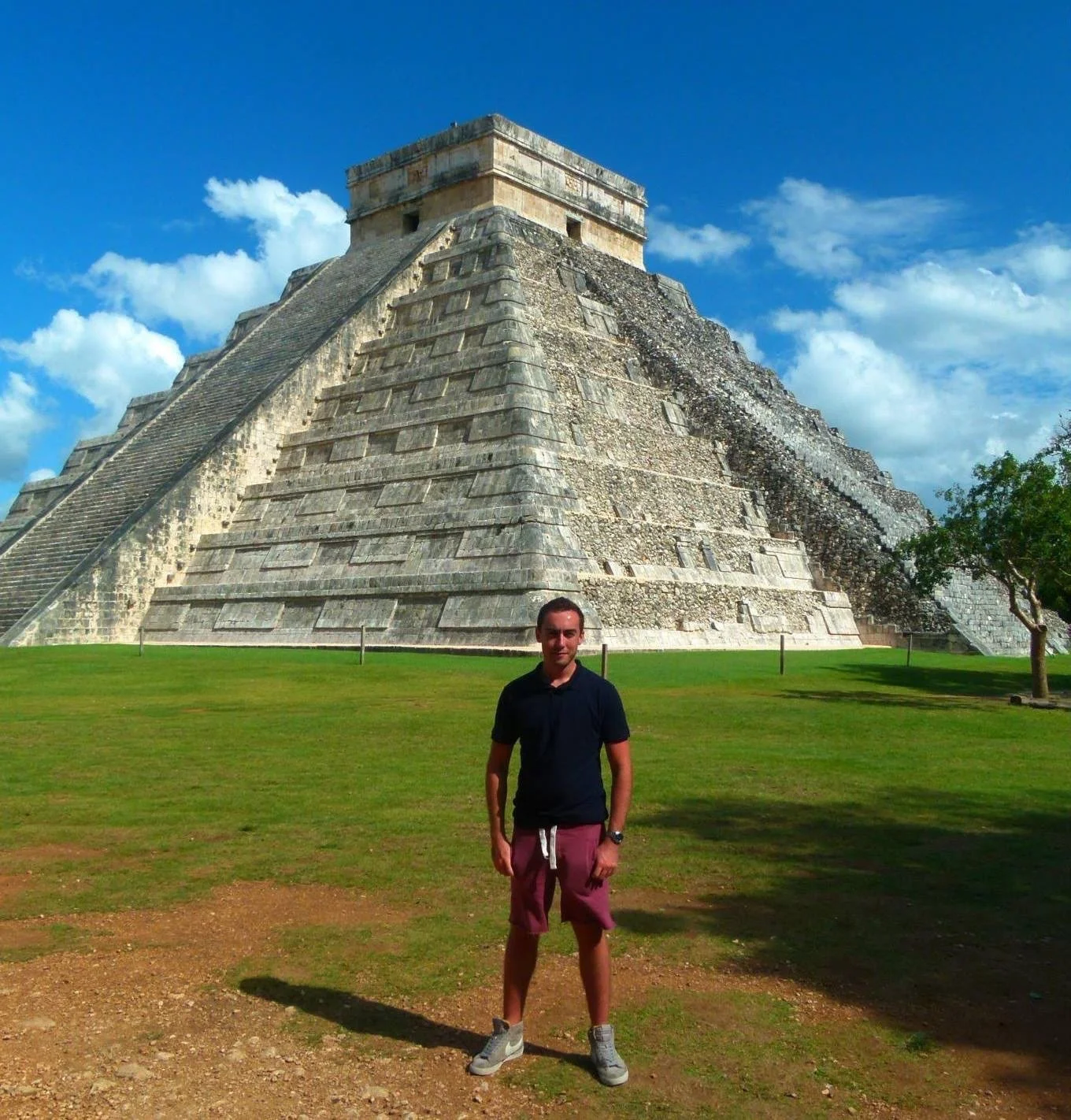 A man standing on grass in front of the ancient Mayan pyramid of El Castillo at Chichen Itza, with a blue sky and some clouds in the background.