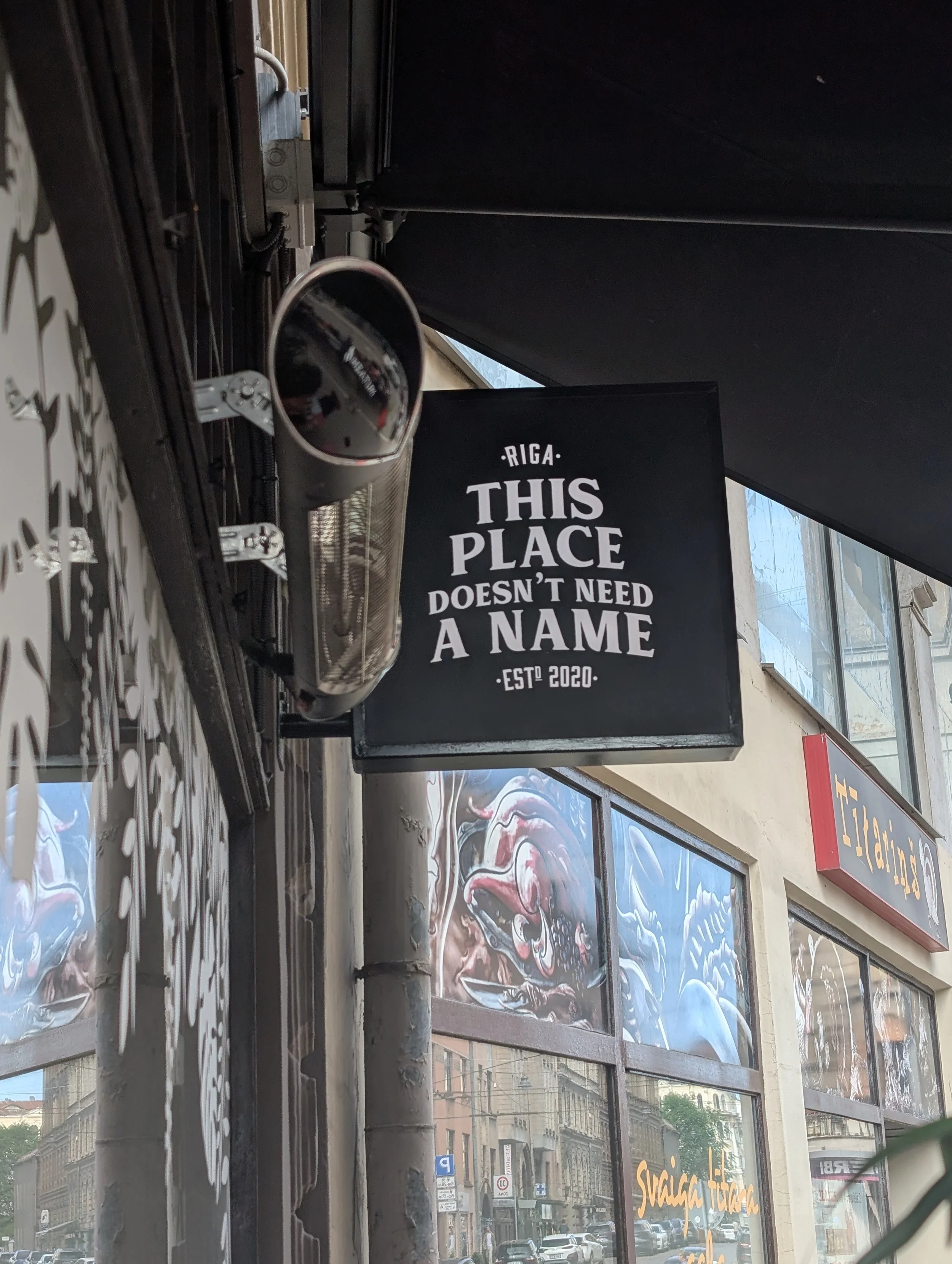 A black sign with white text that reads: 'RIGA THIS PLACE DOESN'T NEED A NAME ESTD 2020'. The sign is hanging outside a building on a city street, with windows and other buildings visible in the background.