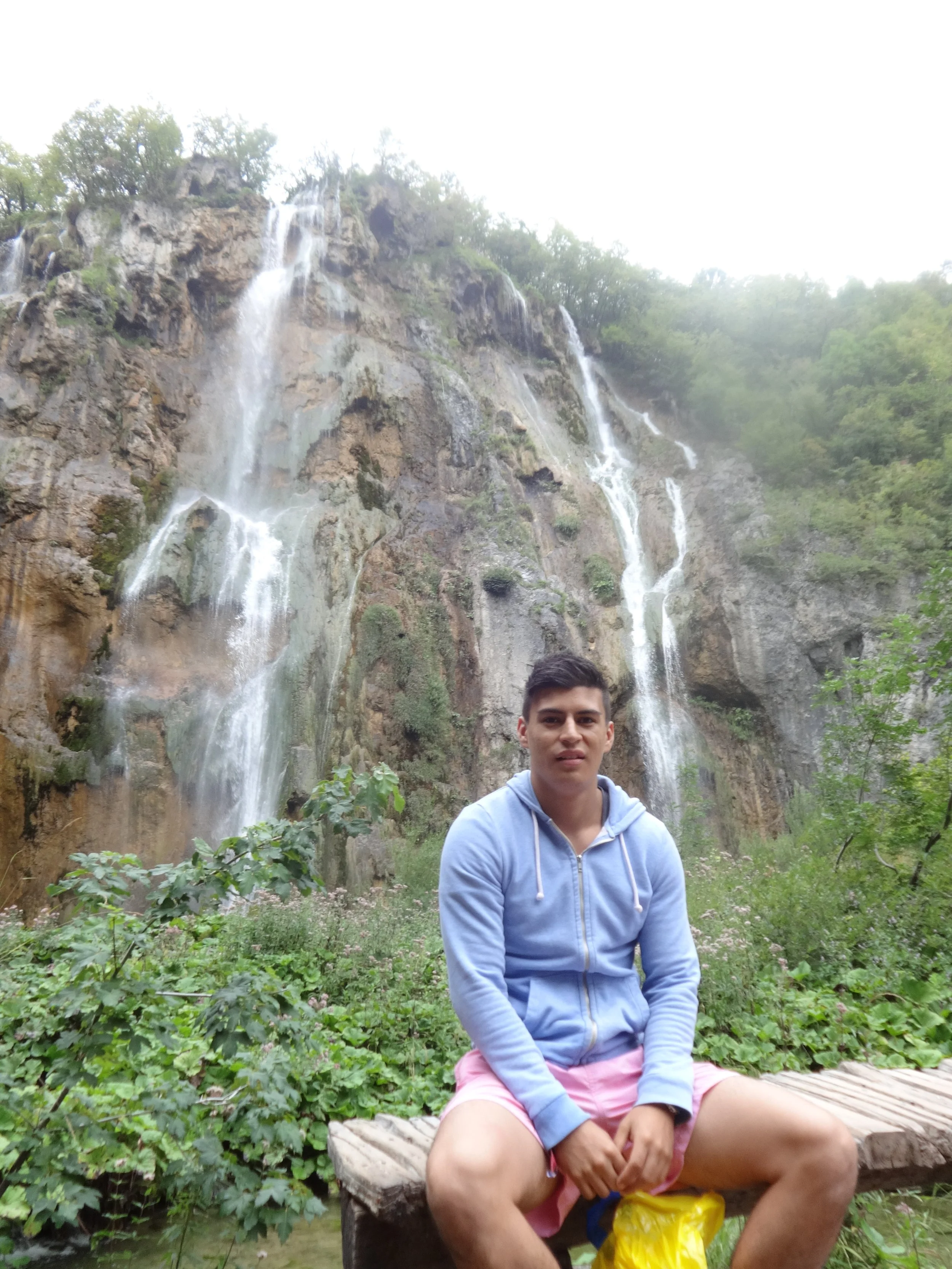 A young man sitting on a wooden bench in front of a tall waterfall surrounded by lush green vegetation.