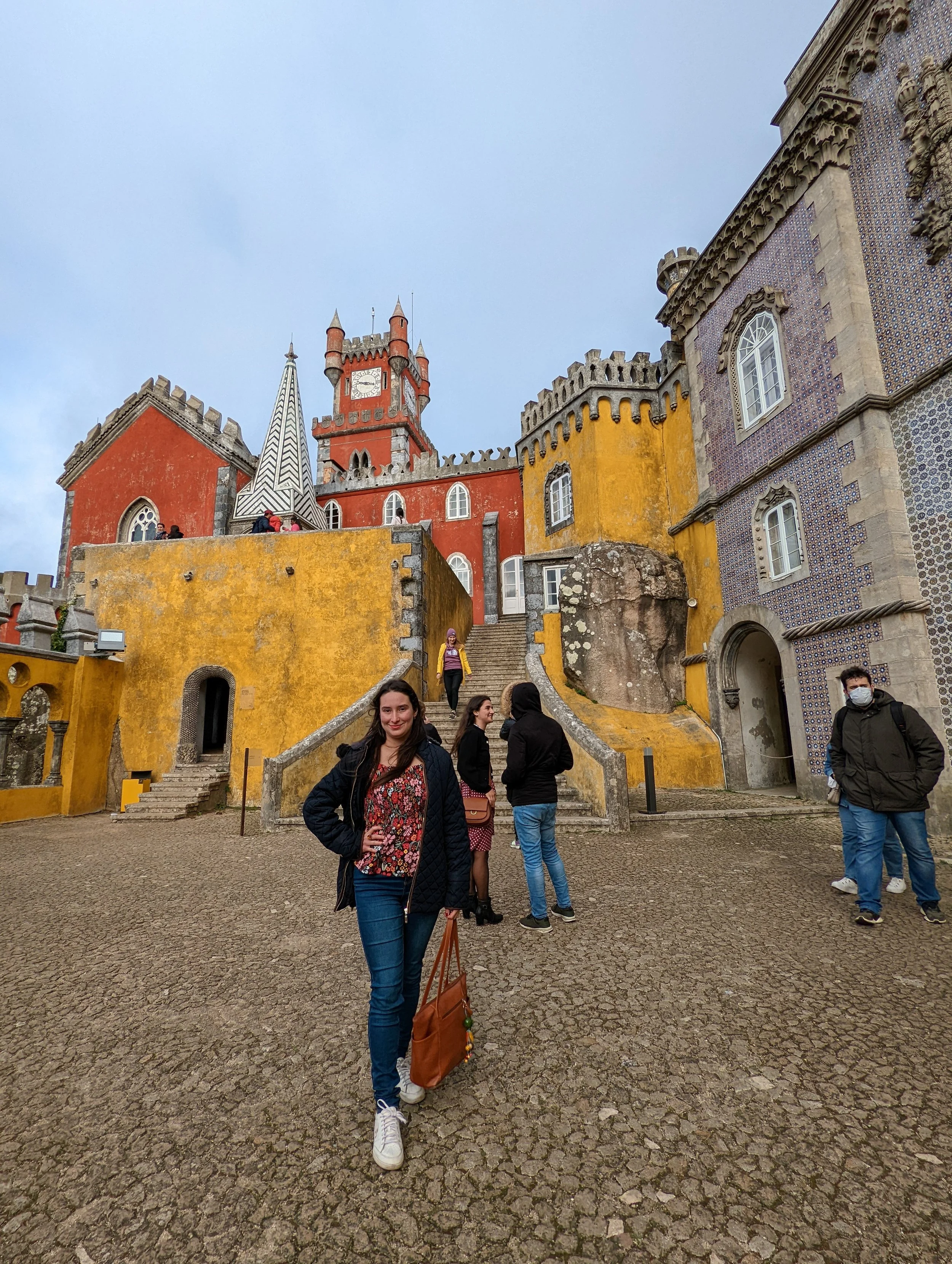 Group of tourists in front of colorful castle with yellow, red, and purple walls, stone stairs, and clock tower, on a cloudy day.