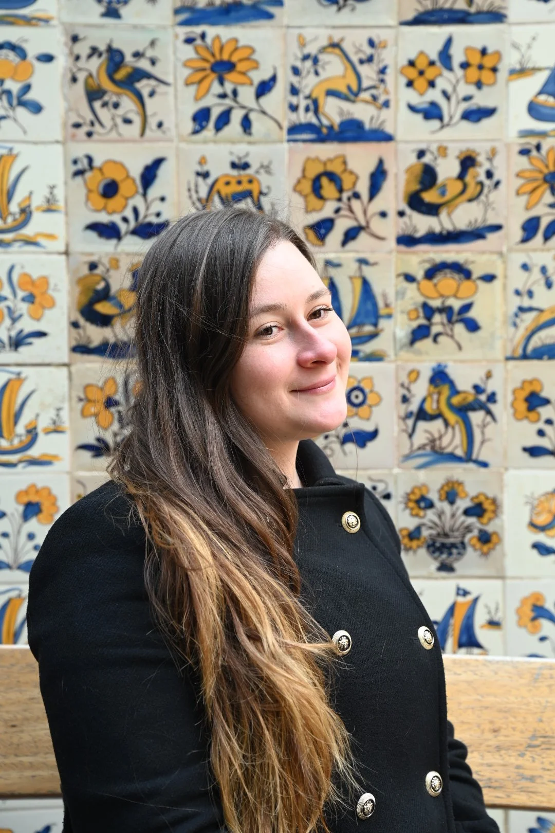 A woman with long brown hair smiling in front of a wall decorated with colorful tiles featuring abstract floral and bird motifs.