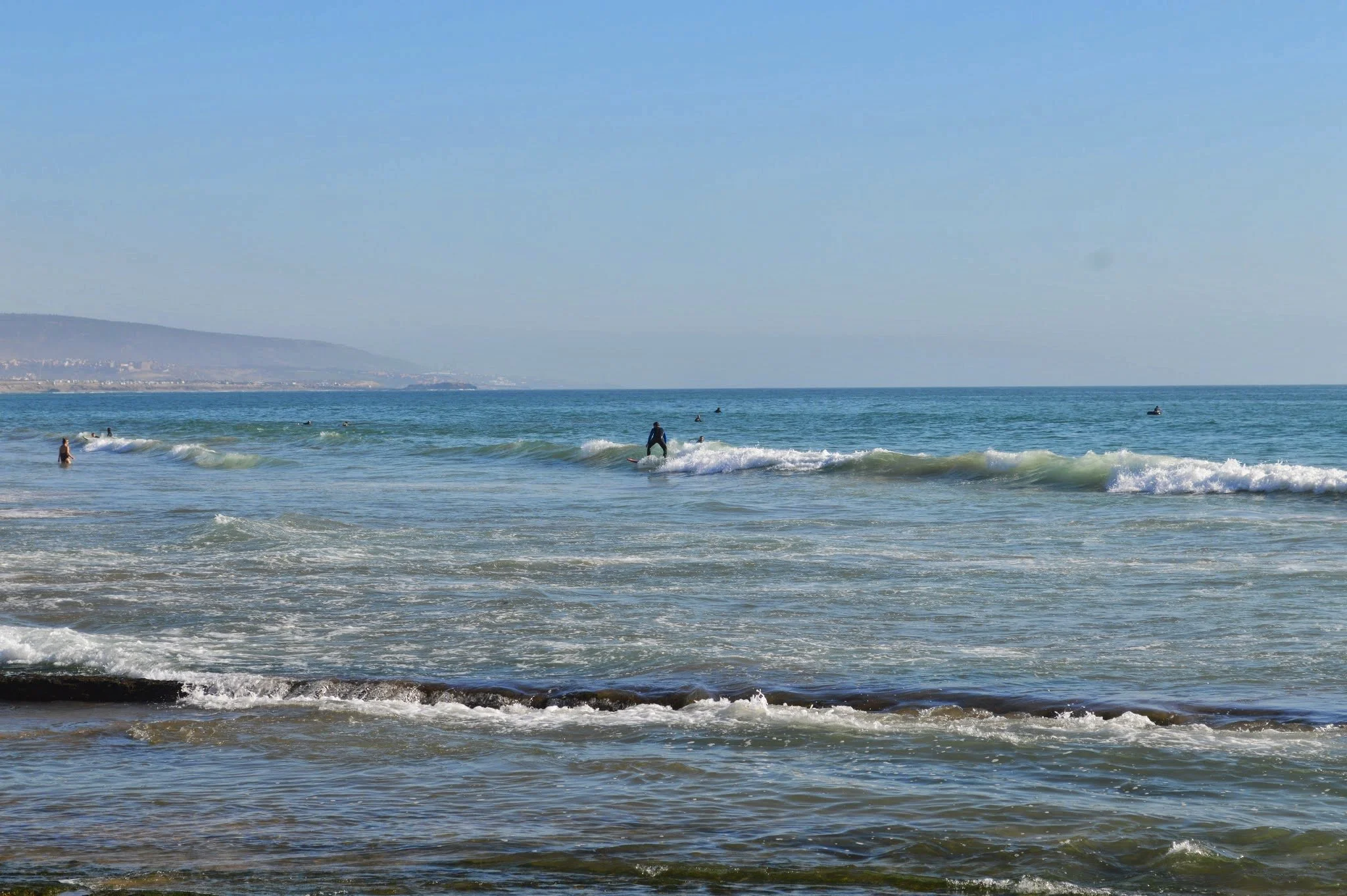 People surfing in the ocean near the shore, with a mountain and a clear sky in the background.