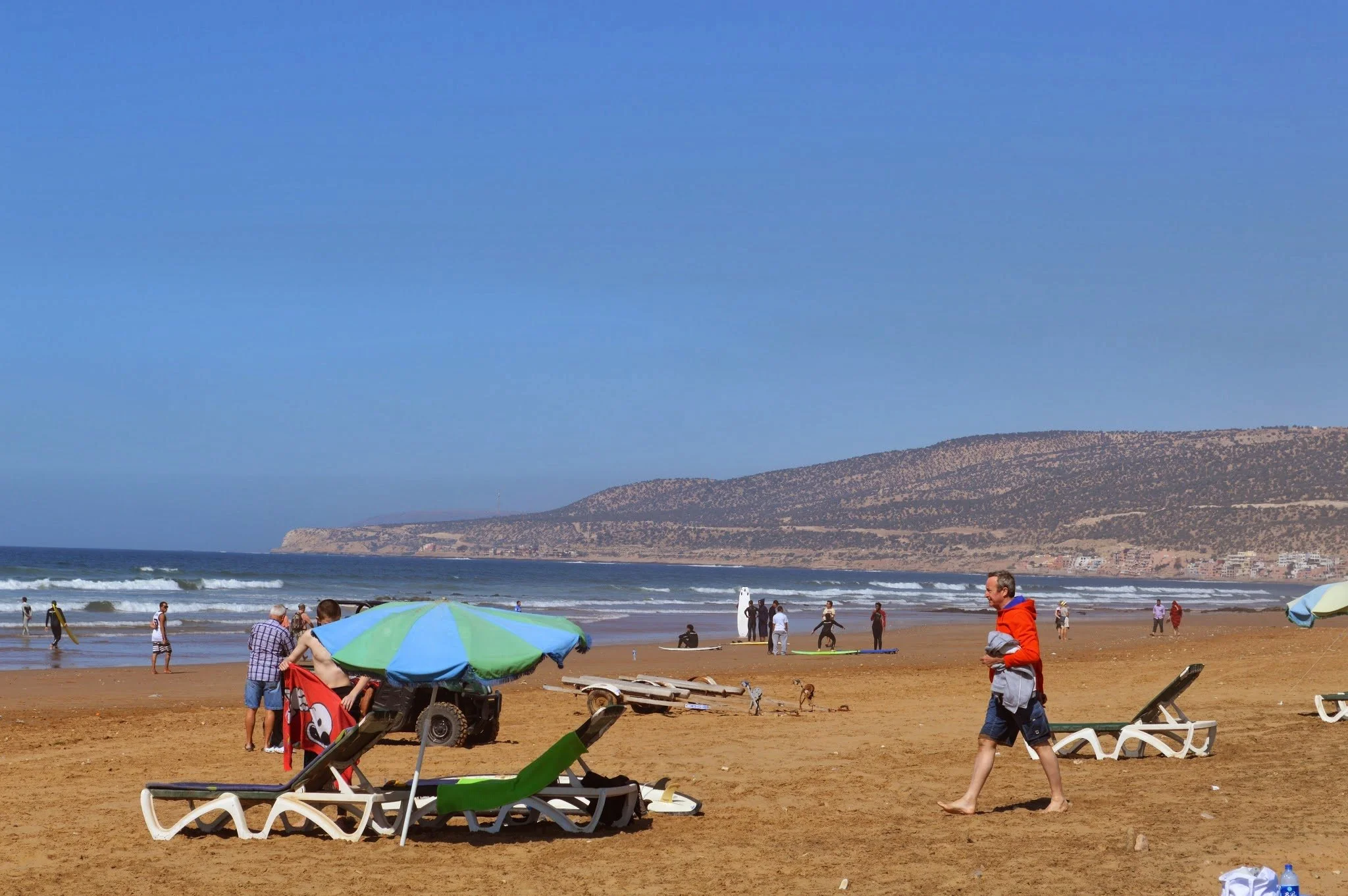 People enjoying a beach day with umbrellas, lounge chairs, and waves in the background. Hills and buildings are visible along the coast.