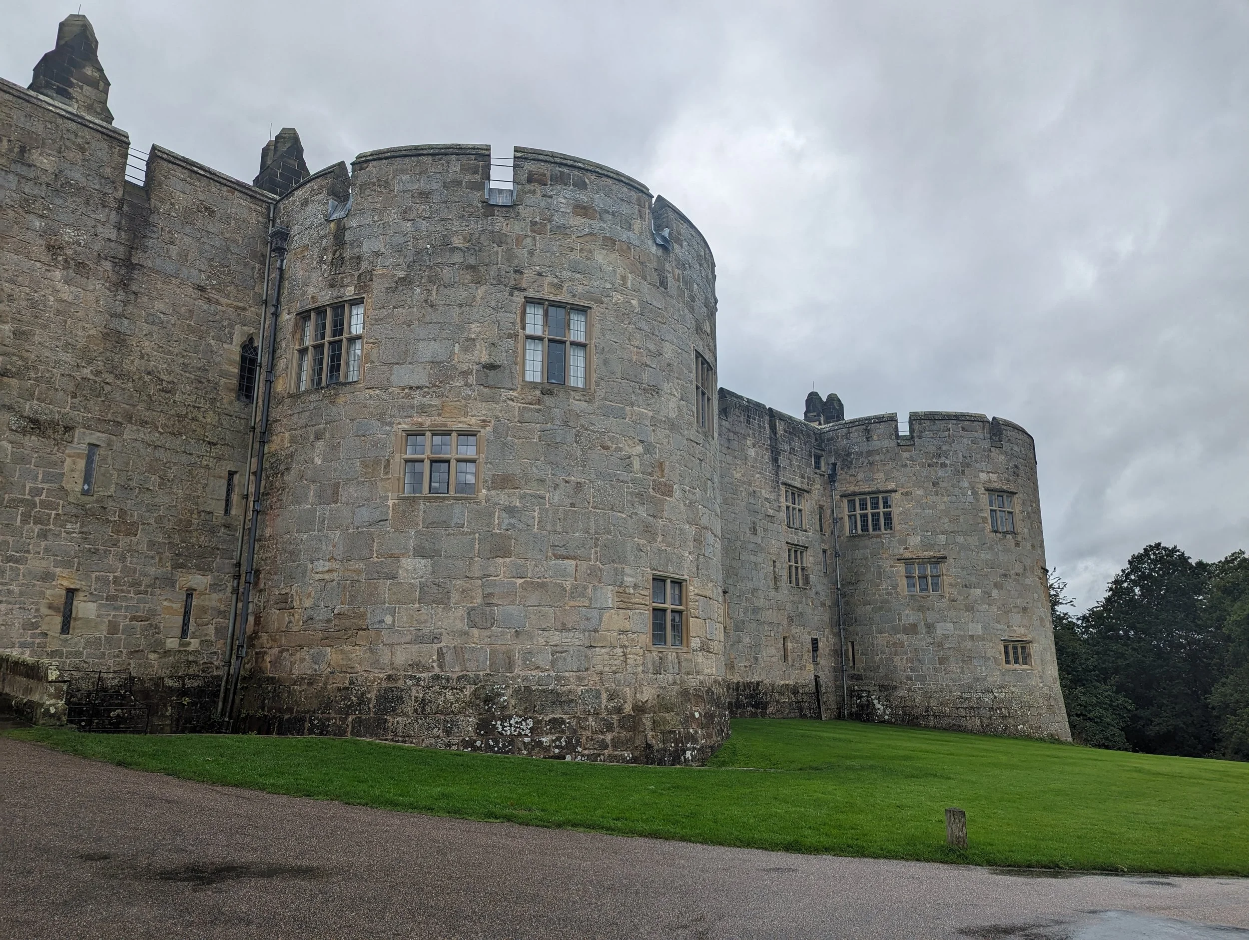 Exterior view of a historic stone castle with rounded towers, multiple windows, and a grassy foreground under a cloudy sky.