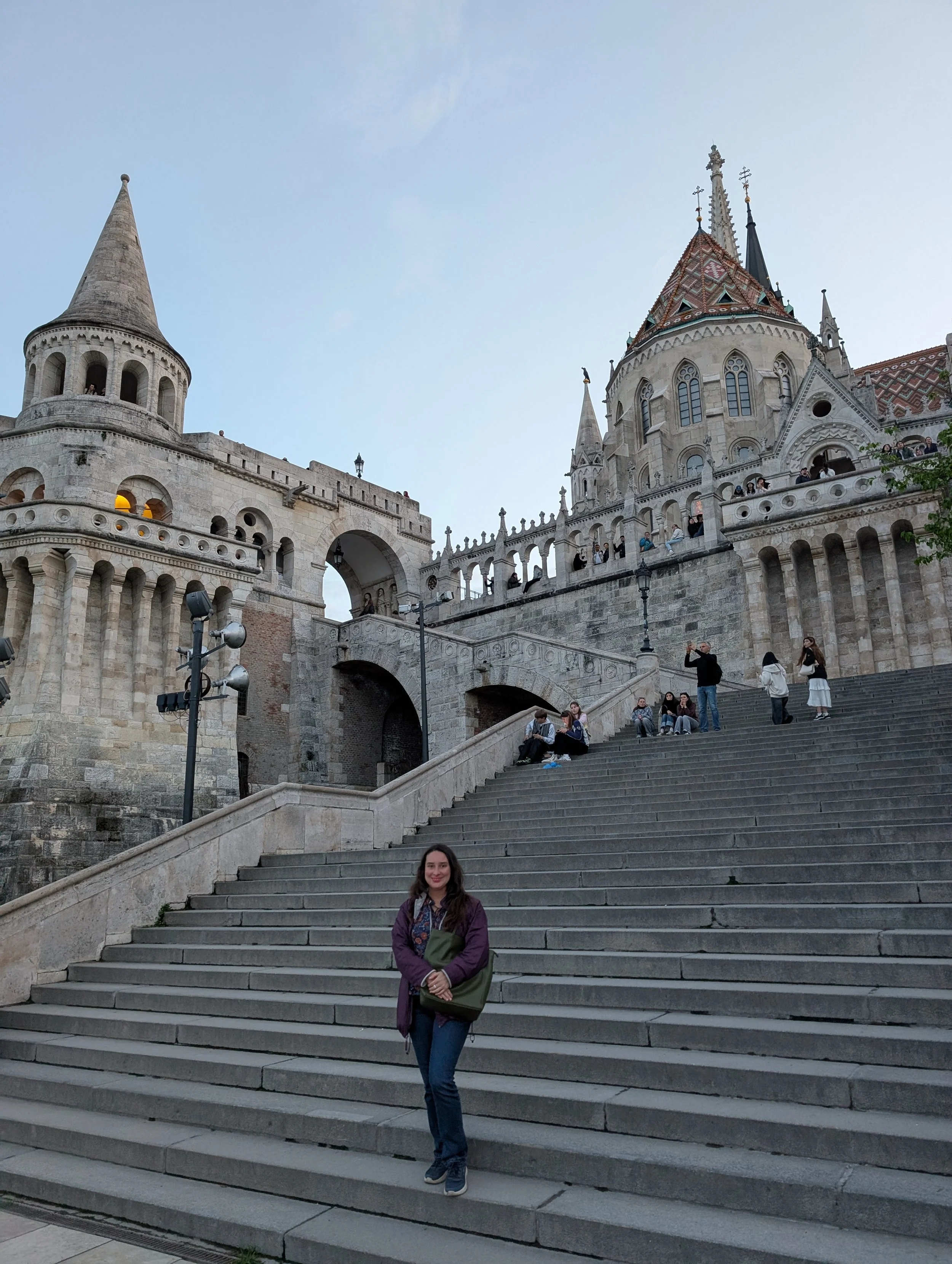 A woman with long dark hair, dressed in a purple jacket and blue jeans, standing on stone steps outside a historic castle with spires and ornate architecture, smiling at the camera.