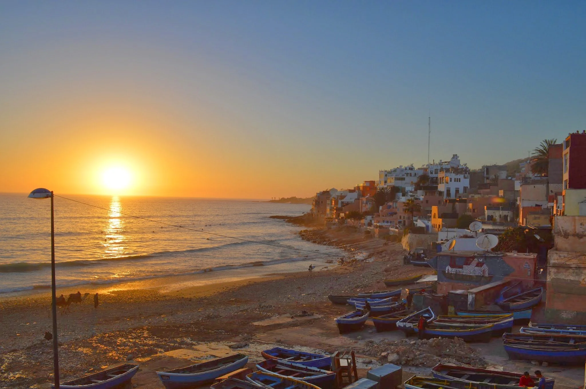 Sunset over a beach with boats on the shore and buildings on a hillside.
