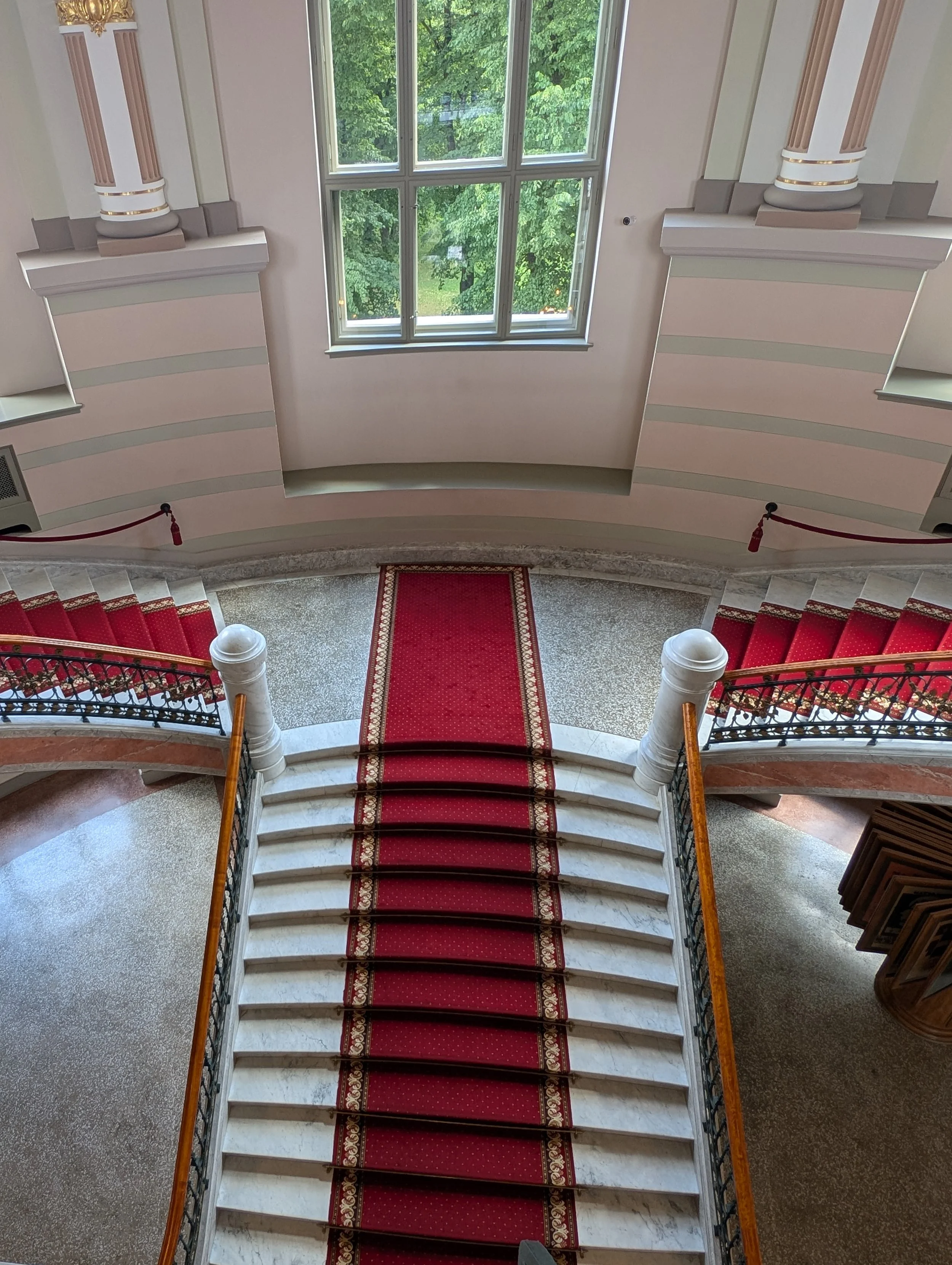 An interior staircase with a red carpet runner, white marble steps, decorative black iron railings, and a view of a large green tree outside a window.