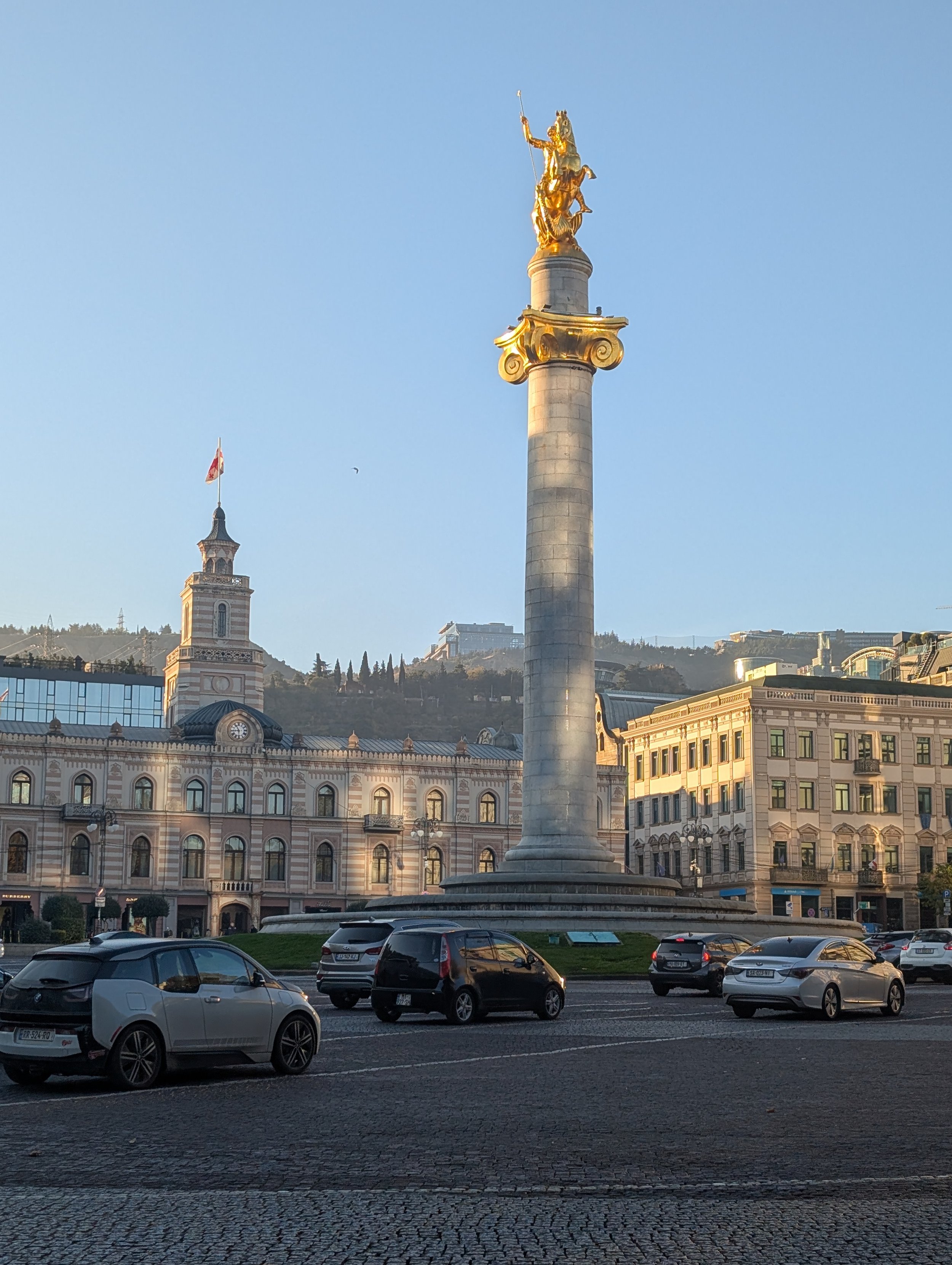 A city square with parked cars in the foreground, historic buildings with ornate architecture and clock towers in the middle ground, and a tall column with a golden statue on top against a clear blue sky in the background.