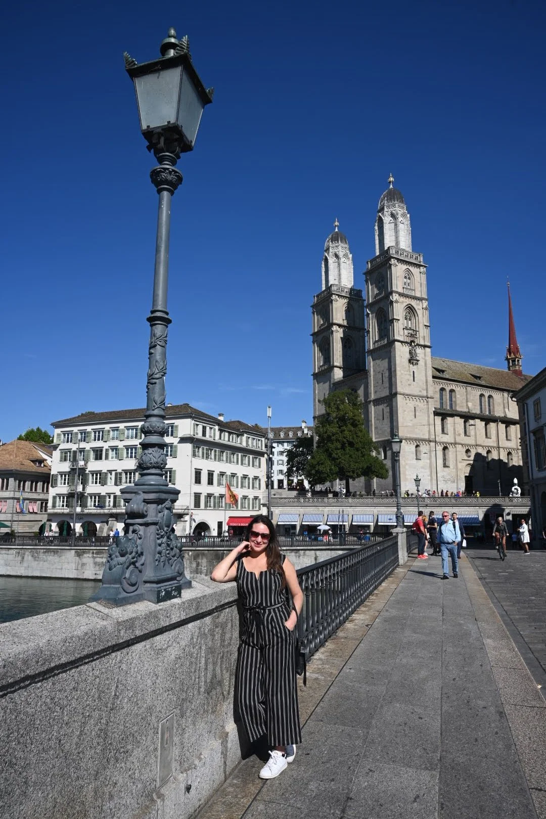 A woman standing on a bridge in front of a large historic church with twin towers in Zurich, Switzerland, on a sunny day.