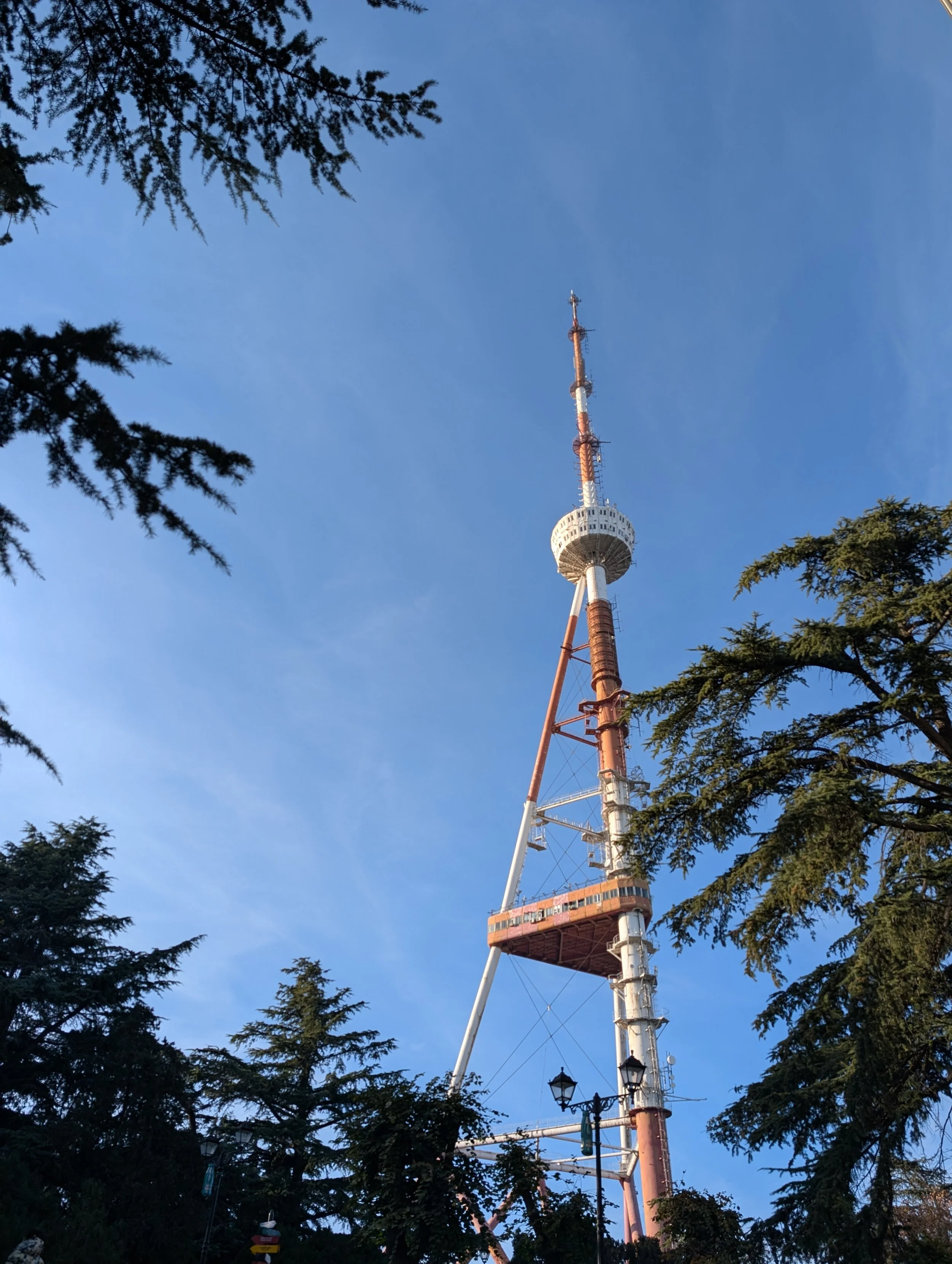 Tall radio or TV tower seen through surrounding trees against a clear blue sky.