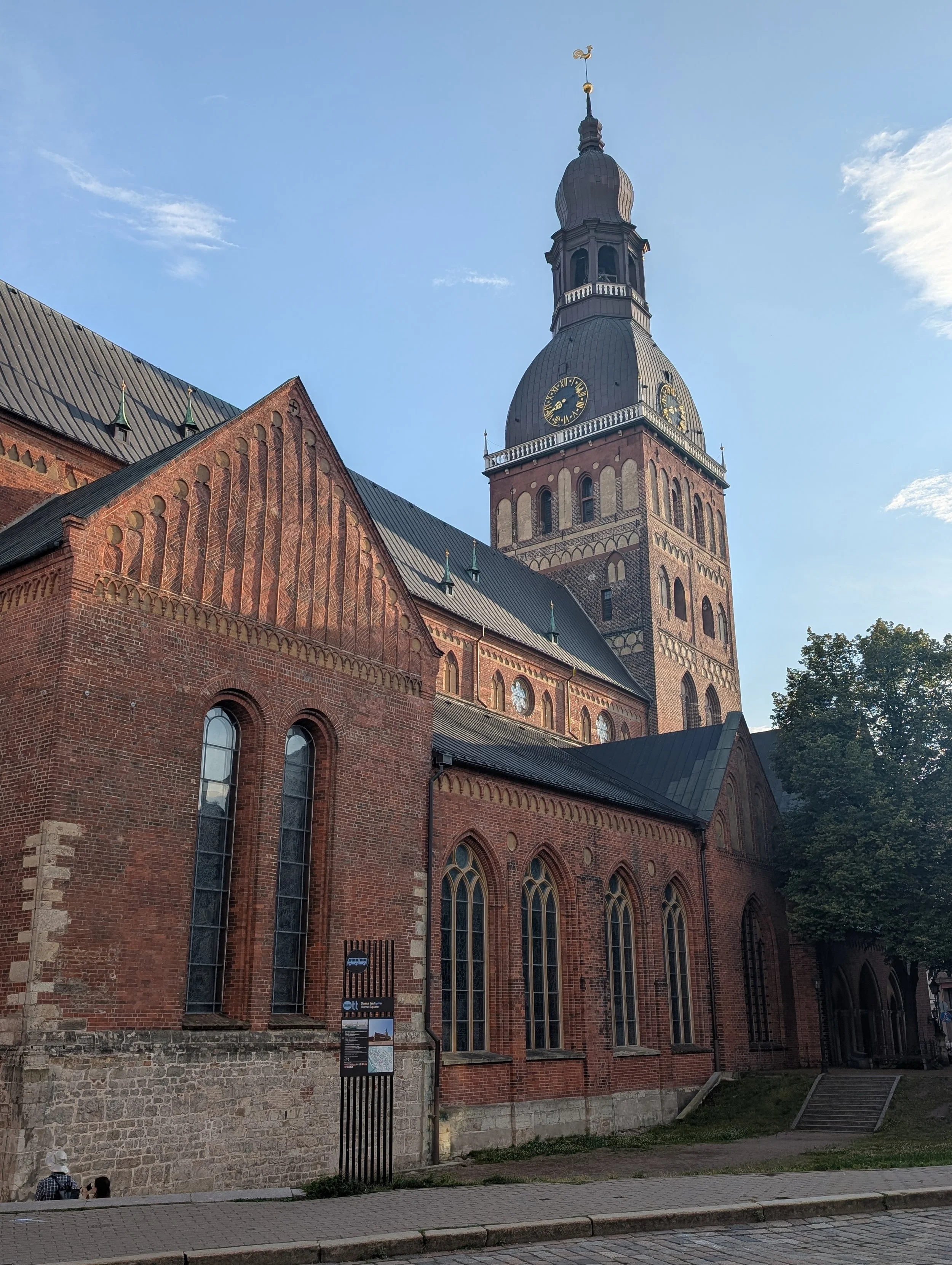 A historic brick church with tall, arched stained glass windows, a clock tower with a domed roof, and a weather vane on top, set against a clear blue sky with some clouds.