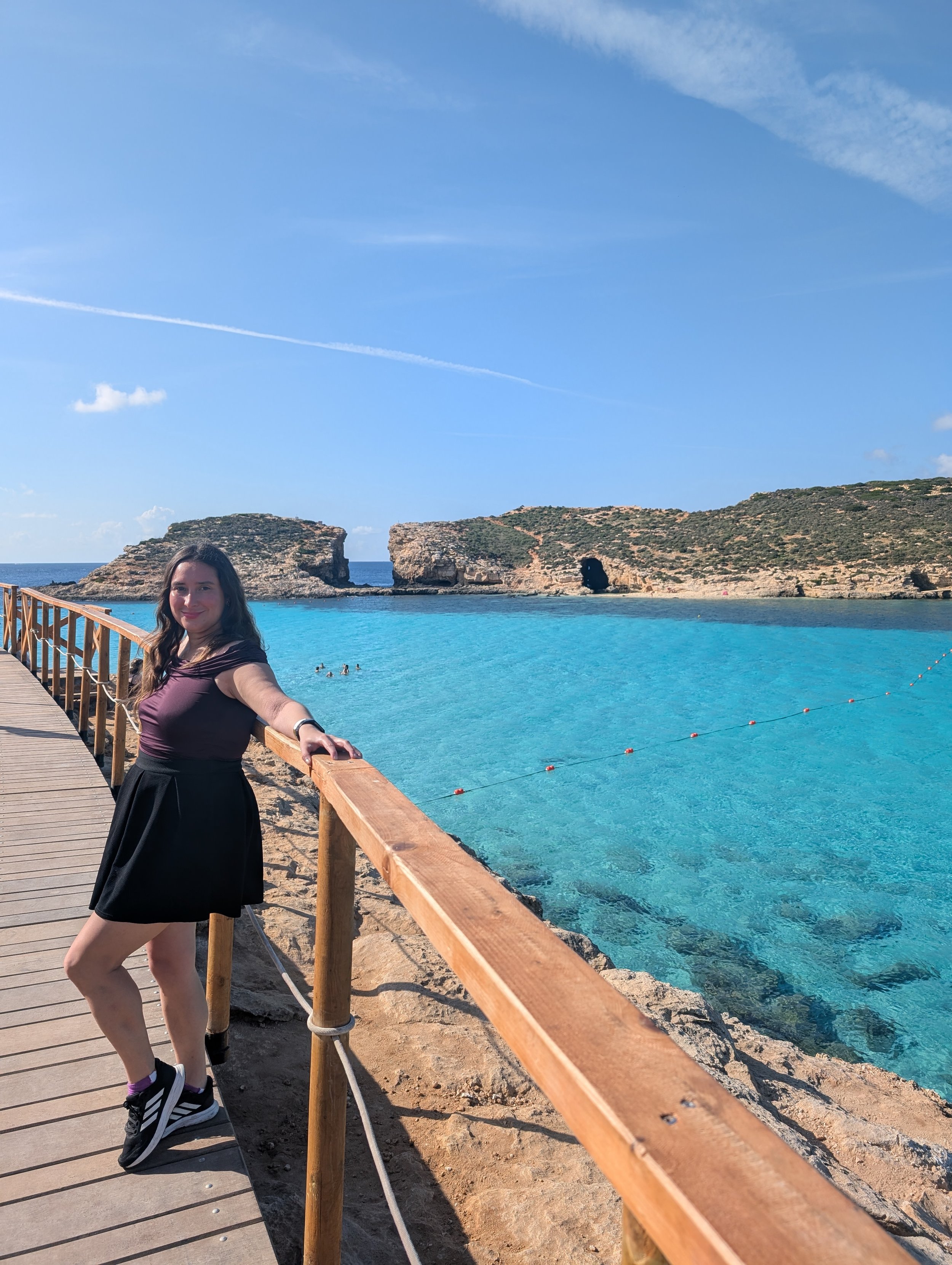 A woman taking a selfie on a wooden pathway by a turquoise beach, with rocky hills in the background under a blue sky with wispy clouds.