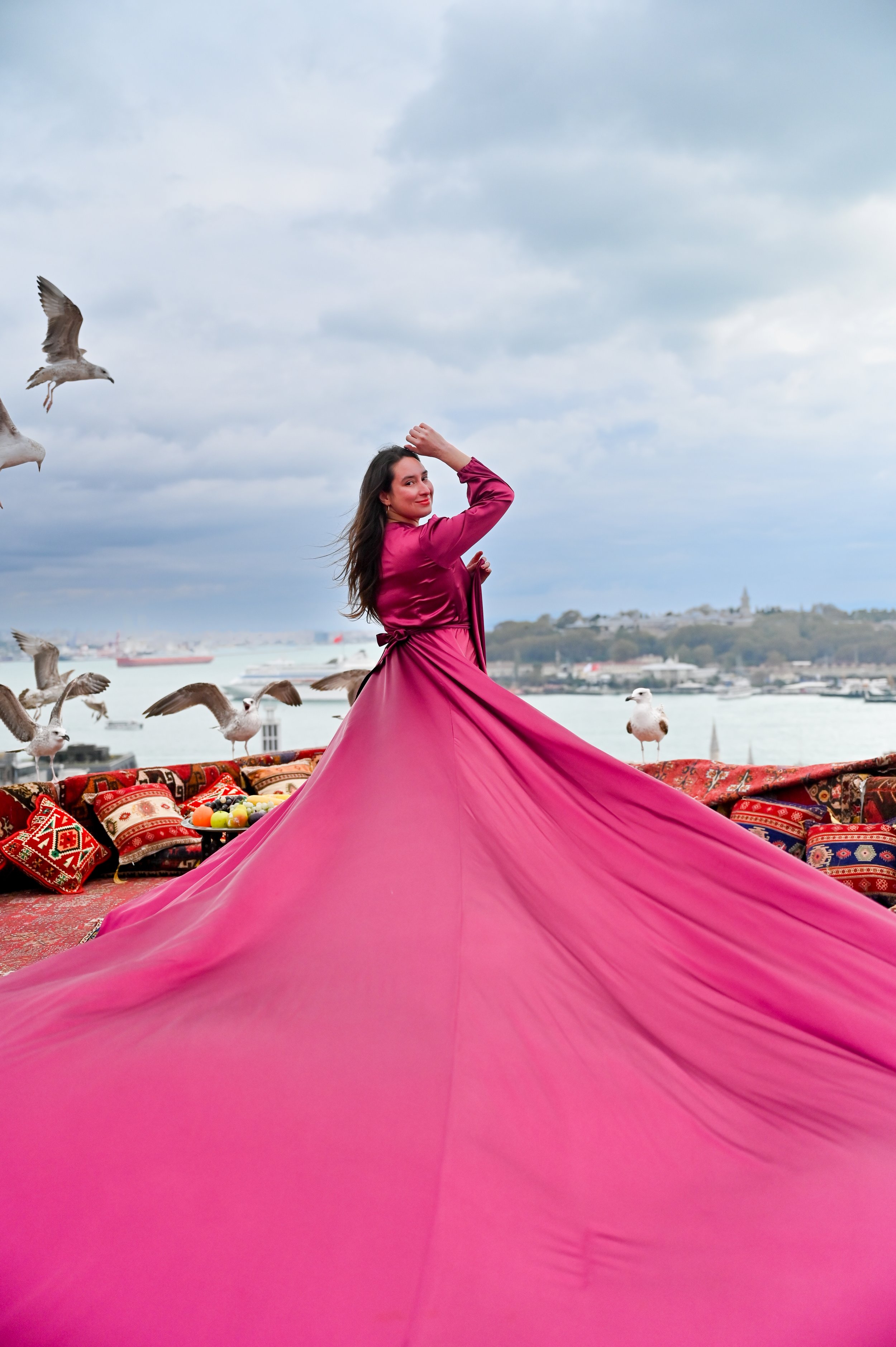Woman in a flowing pink dress standing outdoors near a body of water with seagulls flying around, under a cloudy sky, with a city and a hill in the background.