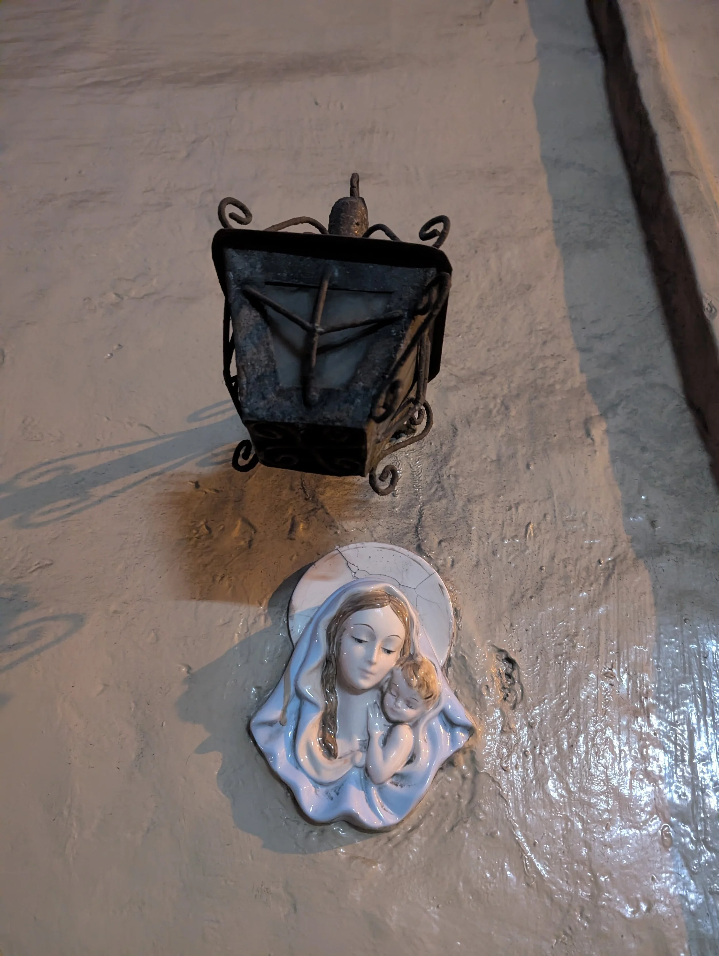 A broken statue or figurine of a woman with a calm, serene expression, lying on a wet surface, with a small child or baby next to her, and a metal lantern or decorative object nearby.