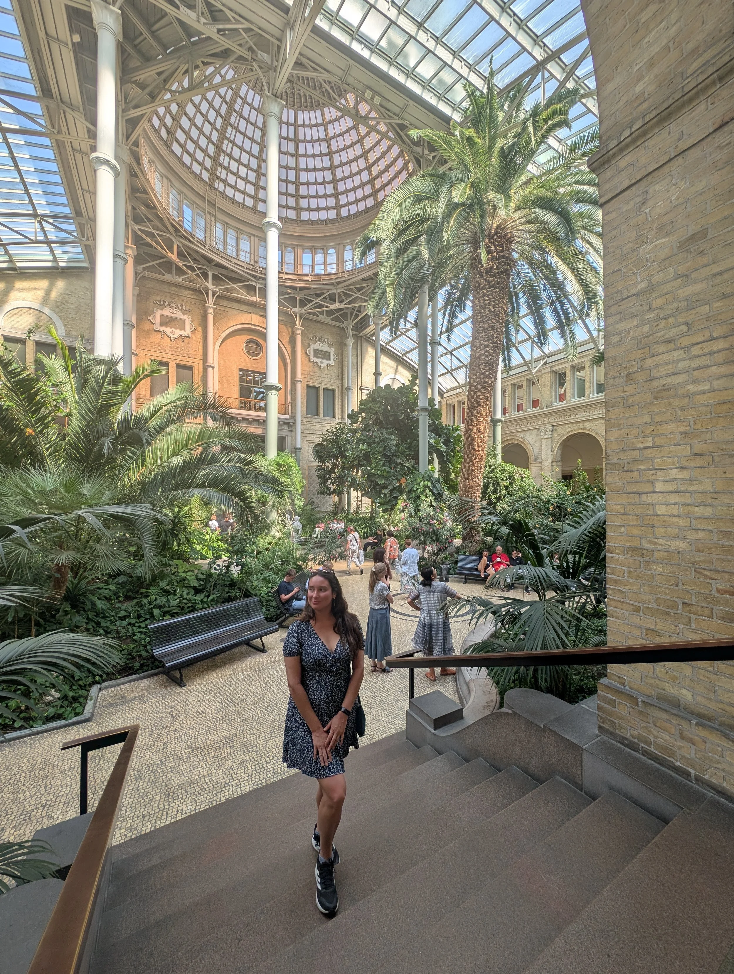 A woman in a blue floral dress with black sneakers walking down stairs inside a glass-roofed conservatory or botanical garden. The background features lush green plants and a large palm tree, with several people enjoying the space.