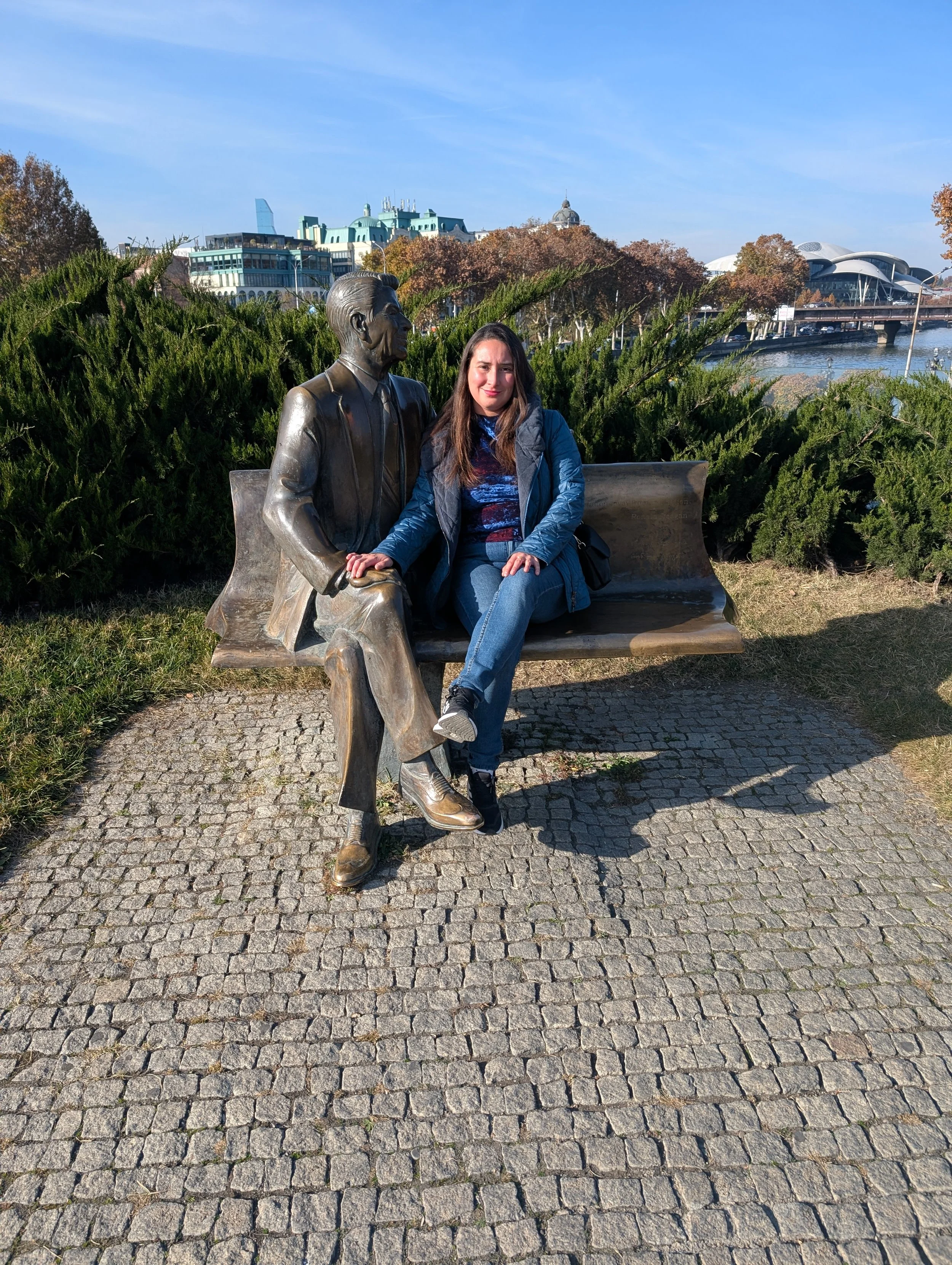 A woman sitting on a park bench next to a bronze statue of a man, with a cityscape and river in the background on a clear, sunny day.