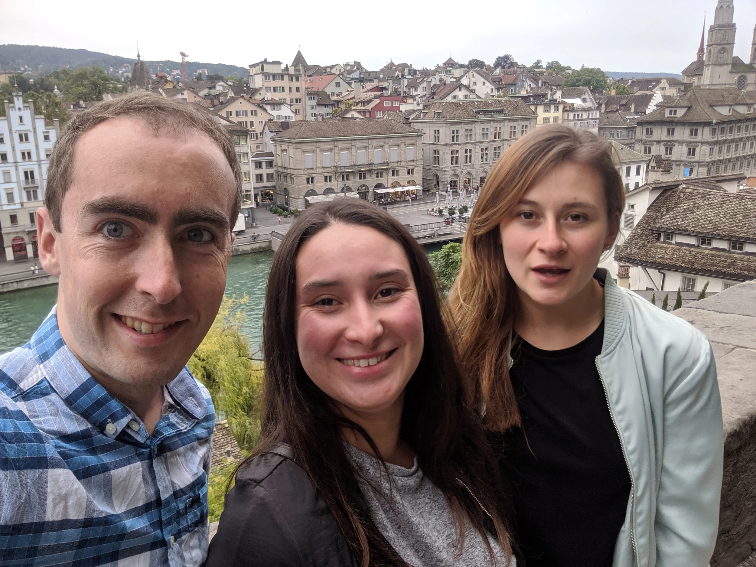 Three people taking a selfie with a European cityscape and river in the background.