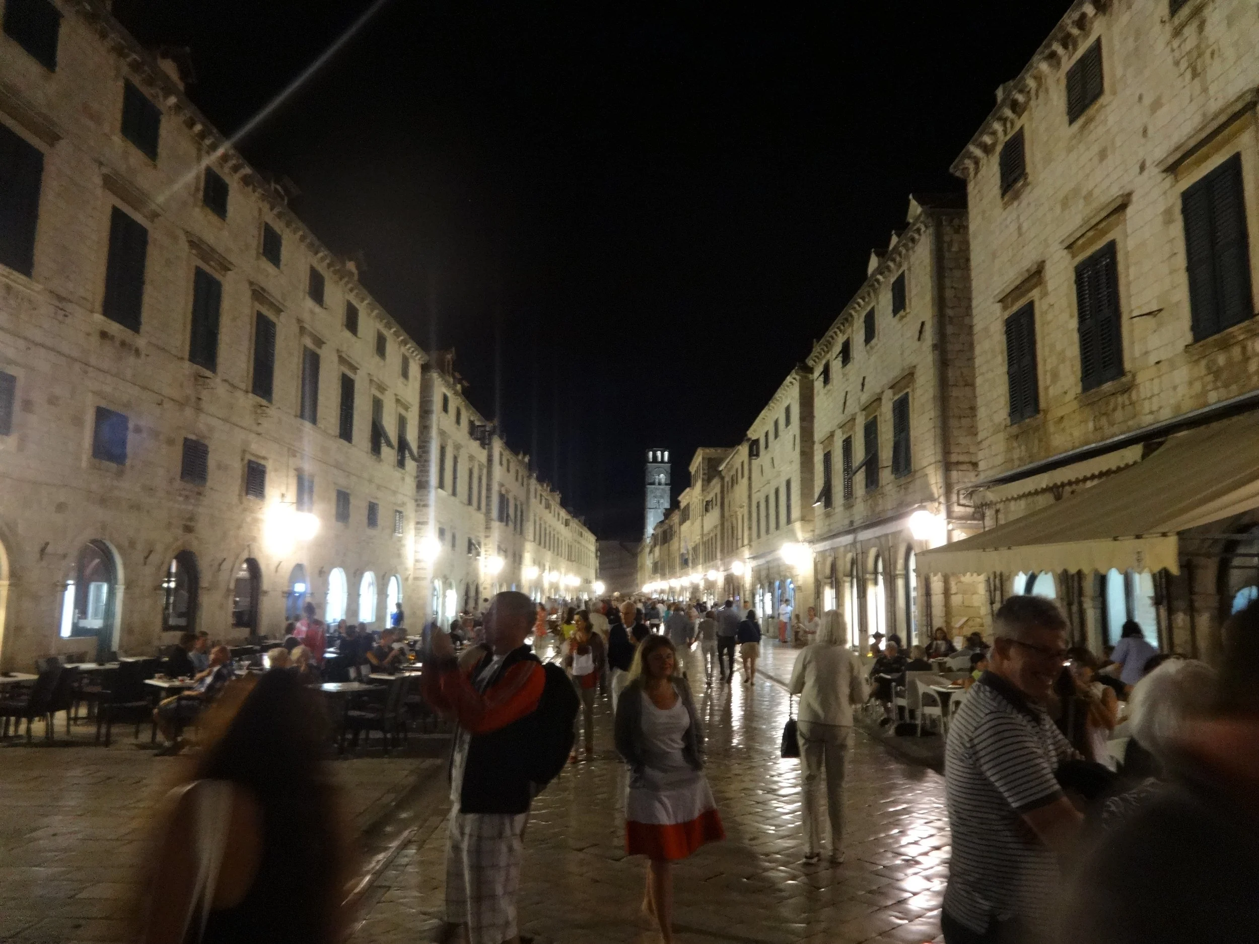 A lively outdoor evening scene with people dining at tables under street lights in a historic city square surrounded by stone buildings with multiple windows and shutters, and a clock tower visible in the background.