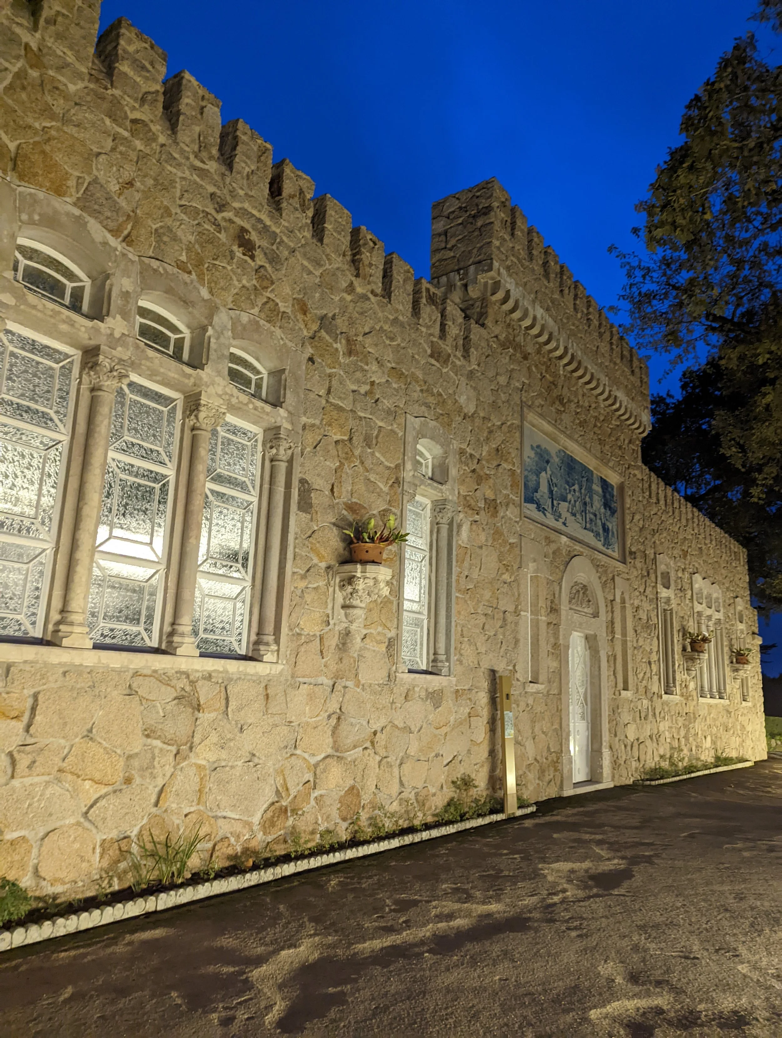 A stone building with decorative windows and wall art, illuminated at night under a dark blue sky, with a tree on the right and a paved walkway in front.