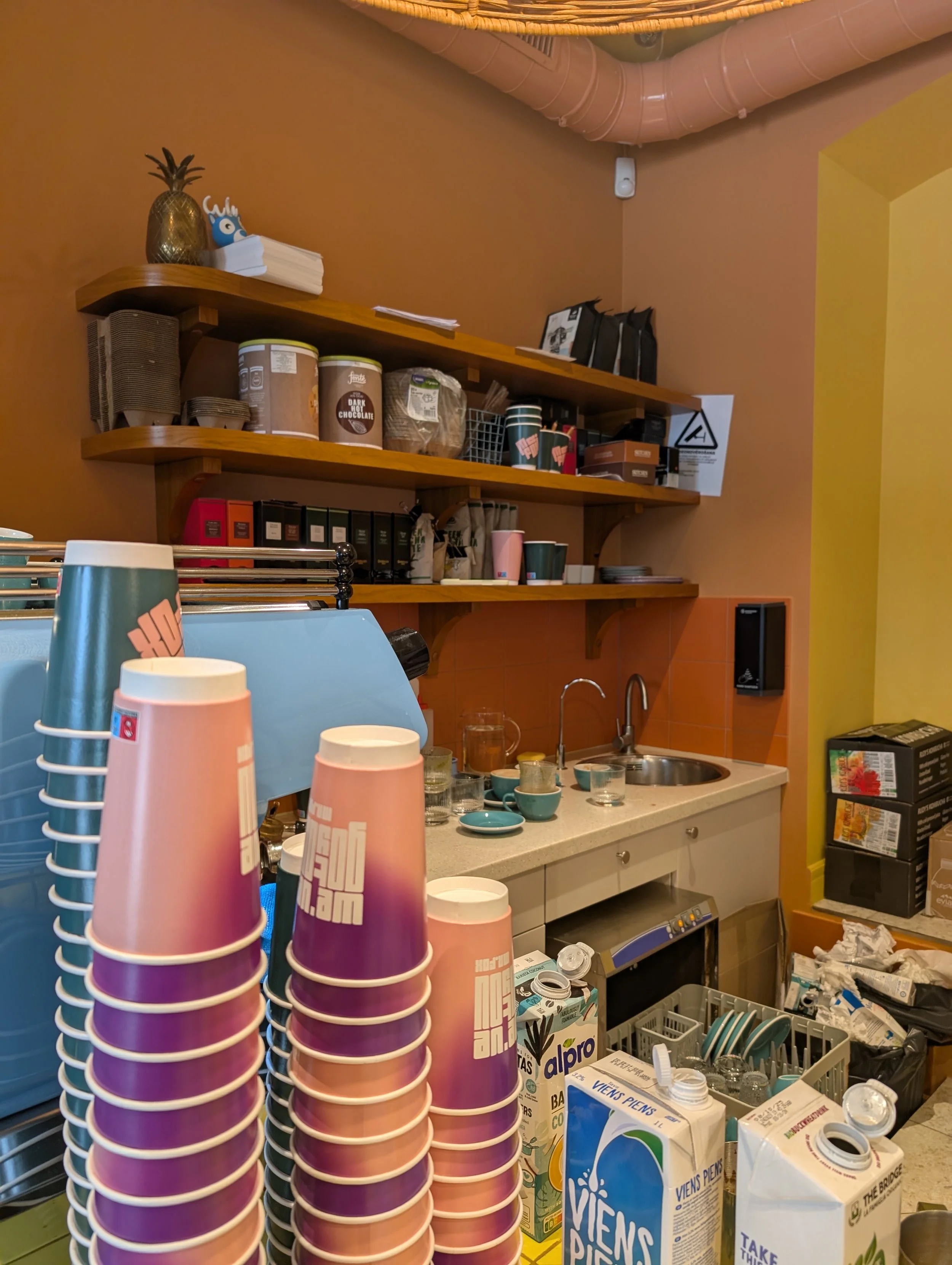 A coffee shop counter with paper cups stacked in various colors in the foreground, and shelves with various supplies like paper cups, containers, and boxes. There are also items like a pineapple-shaped decoration and some napkins on the shelves. The 