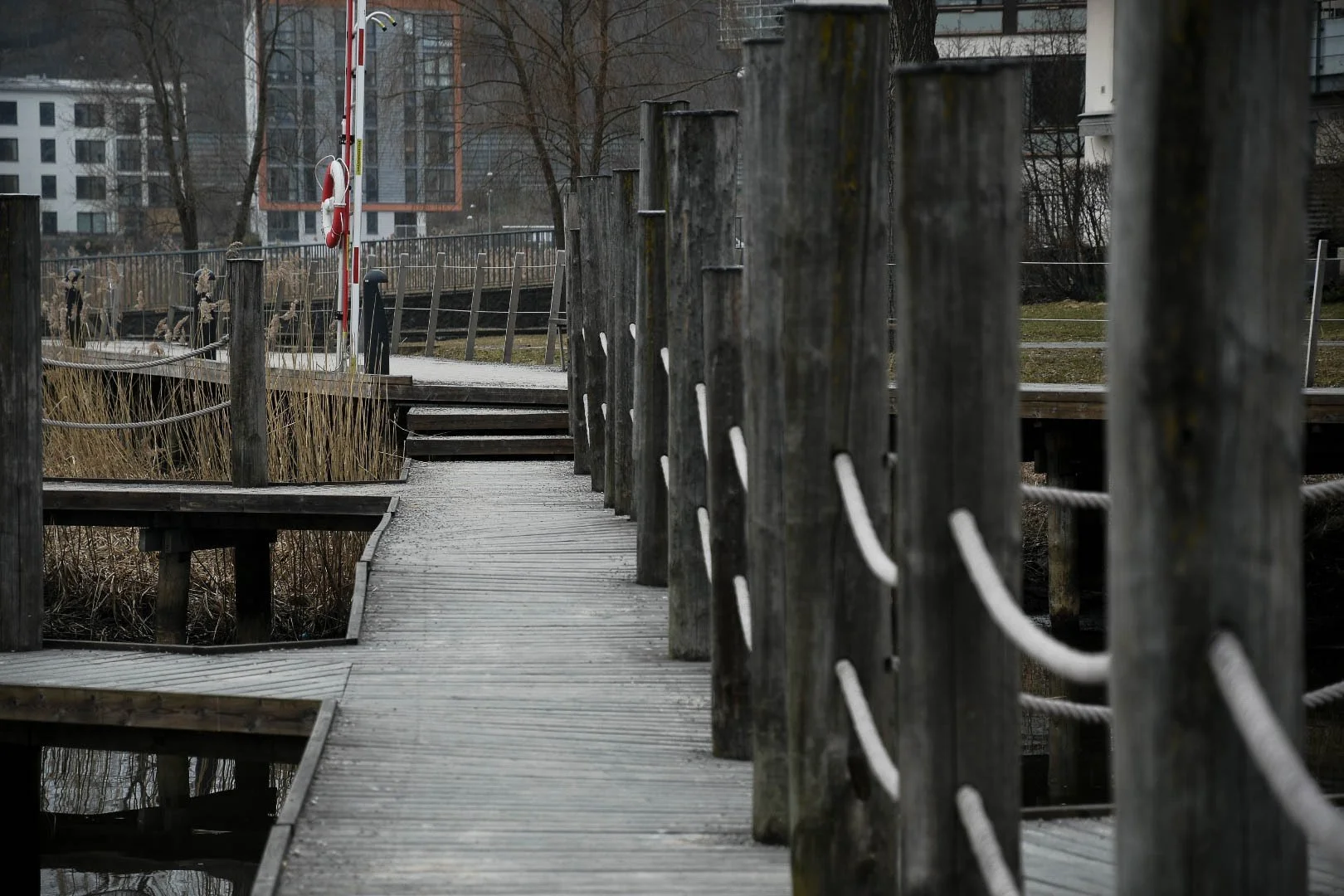 A wooden walkway with a railing over a body of water. In the background, there are buildings and leafless trees on a cloudy day.
