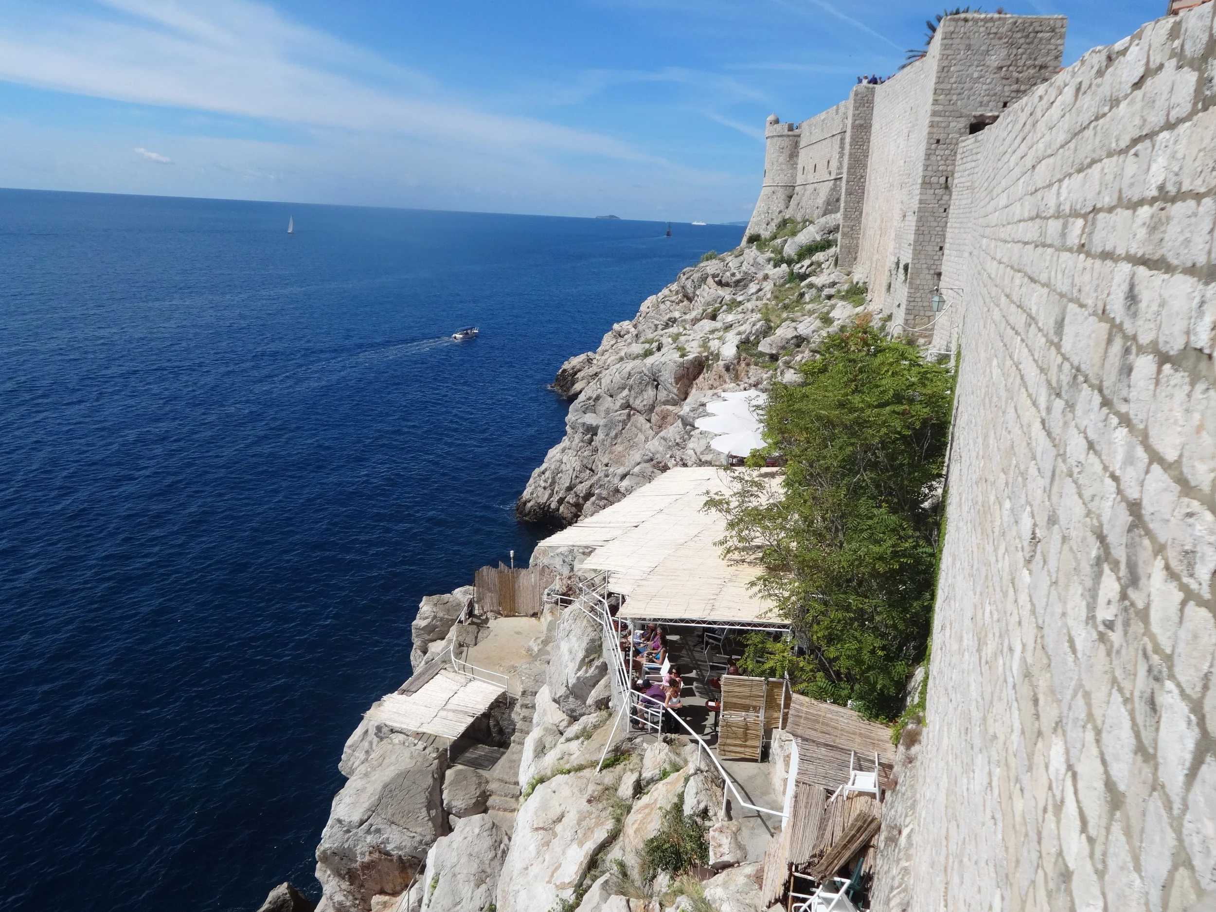 A coastal scene featuring an ancient stone fortress on rocky cliffs overlooking the ocean with a sailing boat and a motorboat on the water, and a clear blue sky overhead.