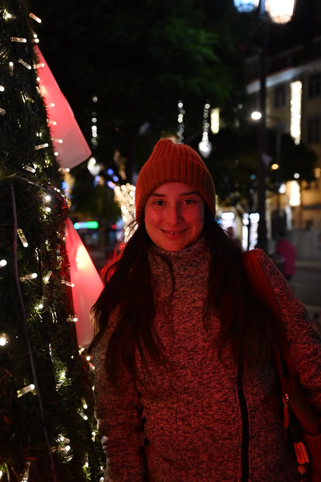 A smiling woman in a red beanie and gray sweater standing next to a Christmas decorated tree with lights, outdoors at night with city lights in the background.