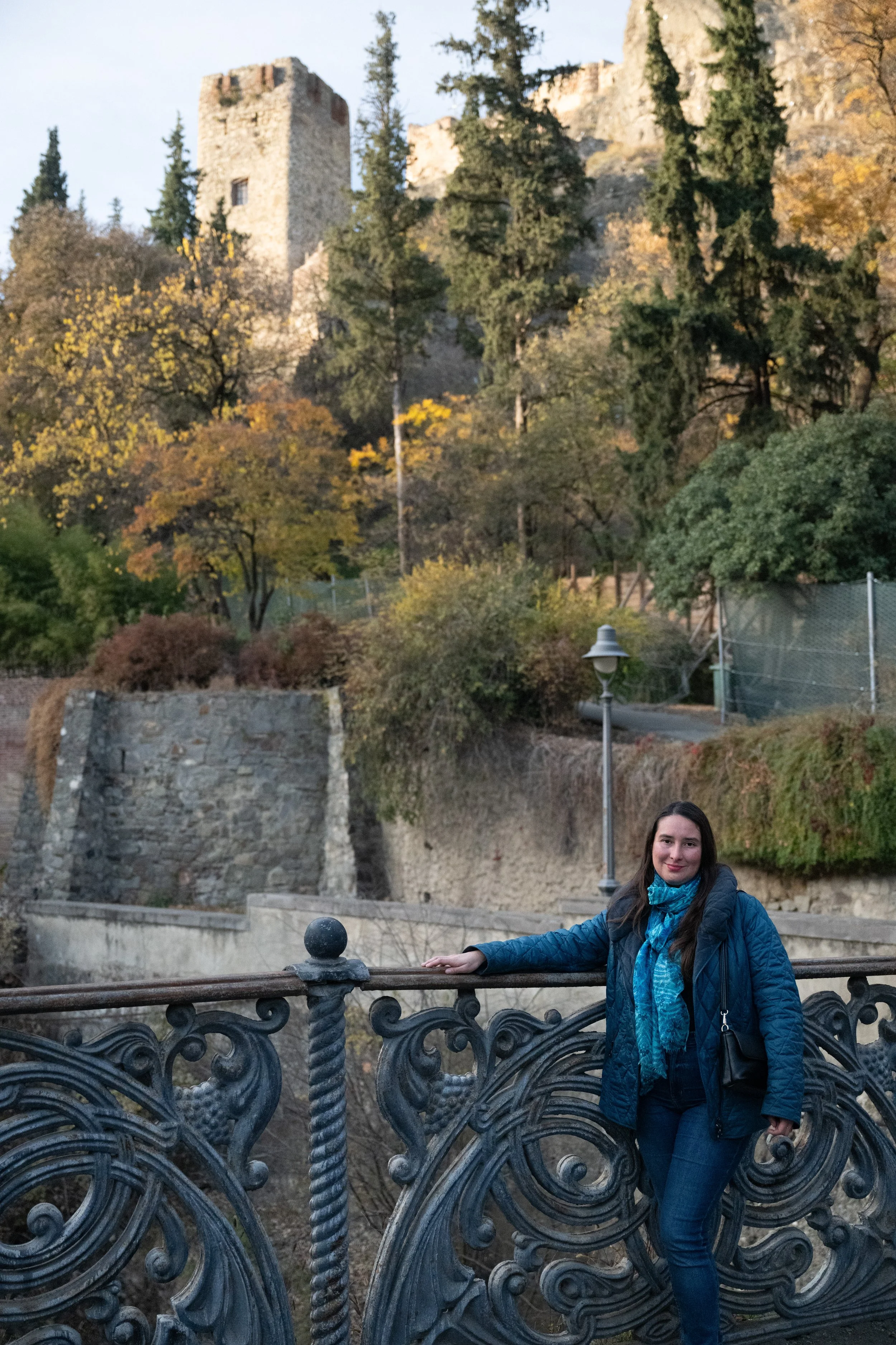 A woman standing on an ornate metal bridge near a river, with a castle and trees with autumn foliage in the background.