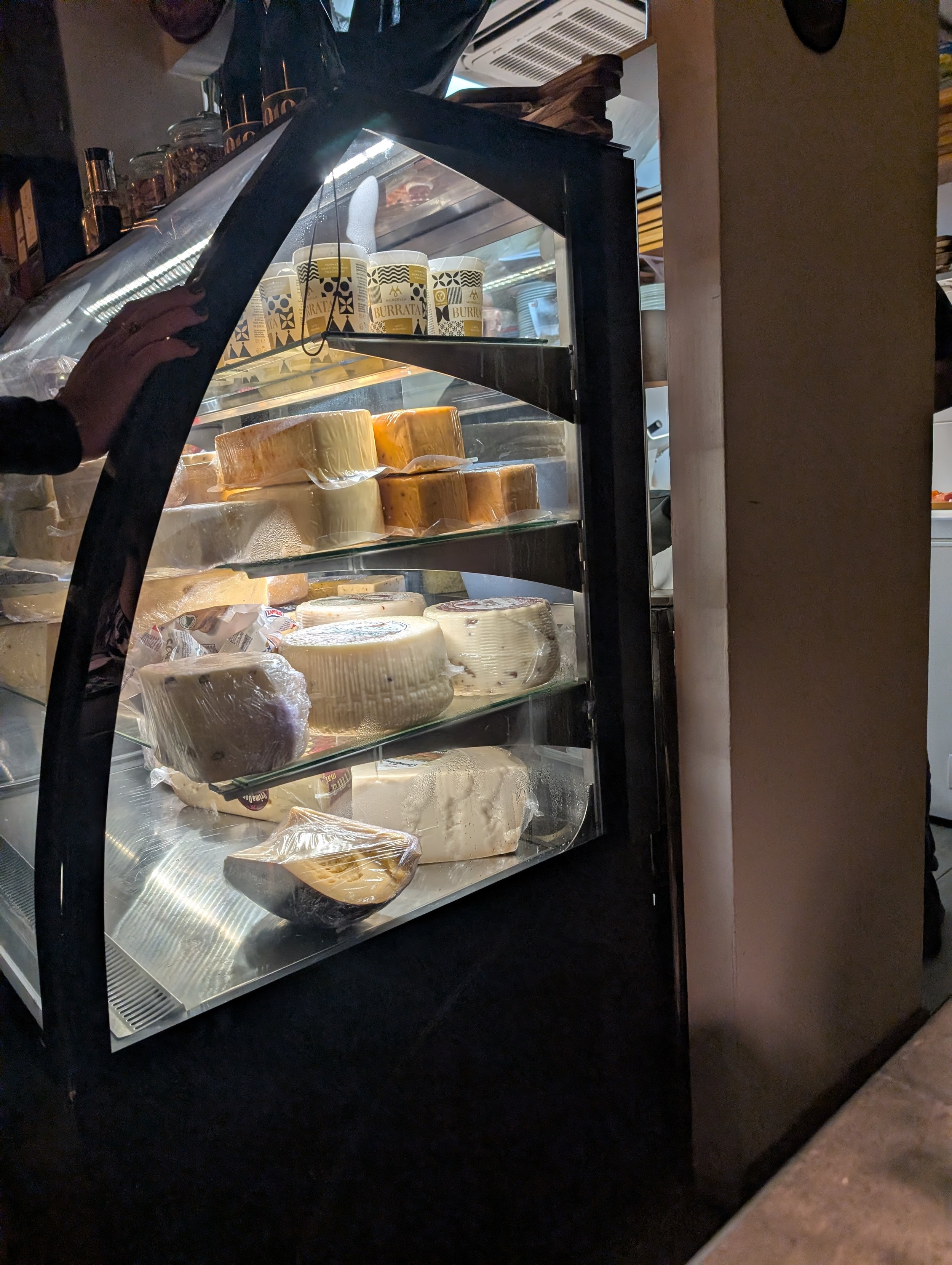 A refrigerated display case filled with various types of cheese, with some wrapped in plastic, in a grocery store or deli. The case has glass doors and is illuminated inside. A hand is reaching towards the cheese, and shelves with jars and other item