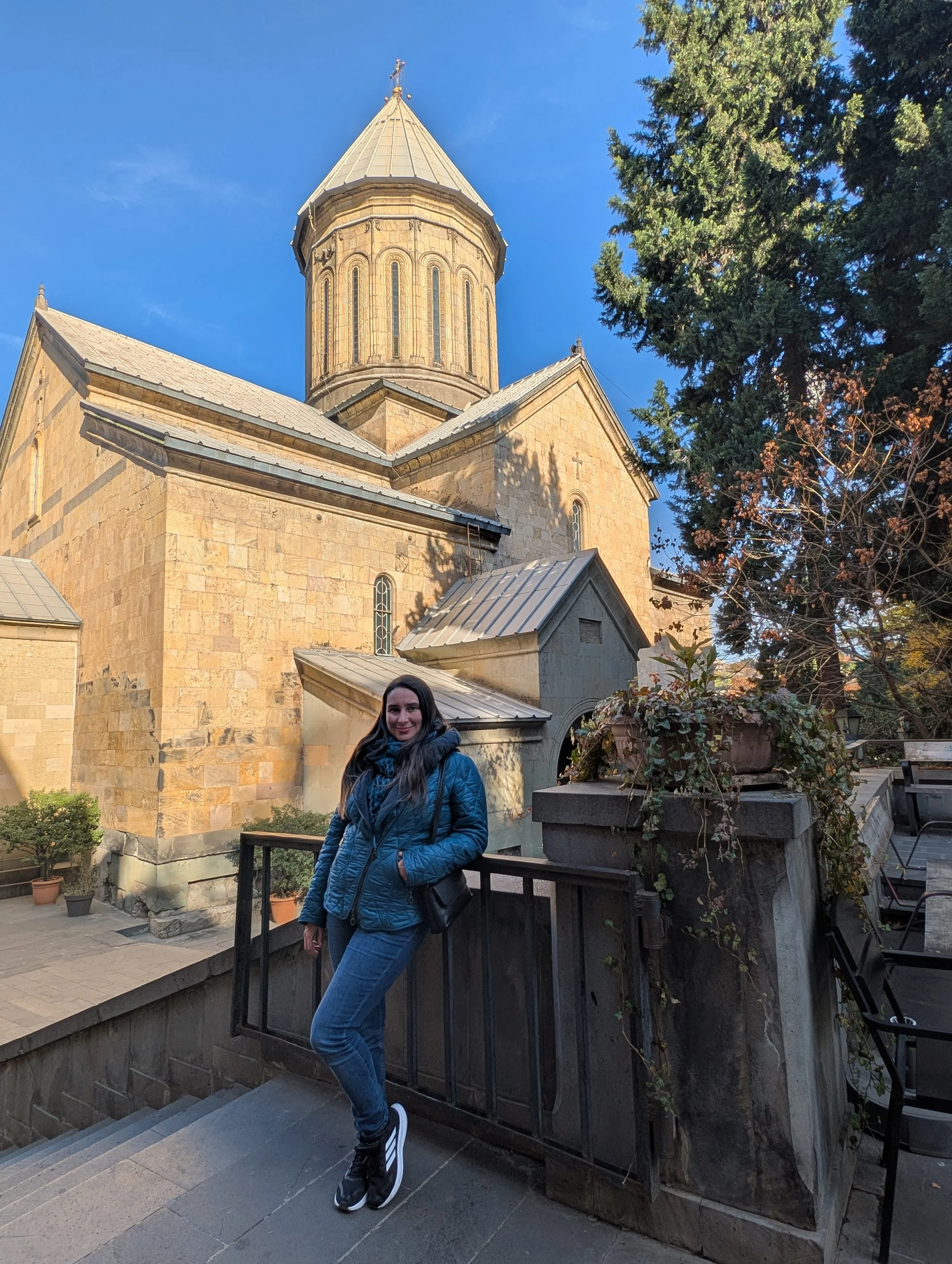 Young woman with long dark hair, wearing a blue jacket and jeans, standing on stone steps outdoors near a stone church with a tall tower, trees, and a clear blue sky.